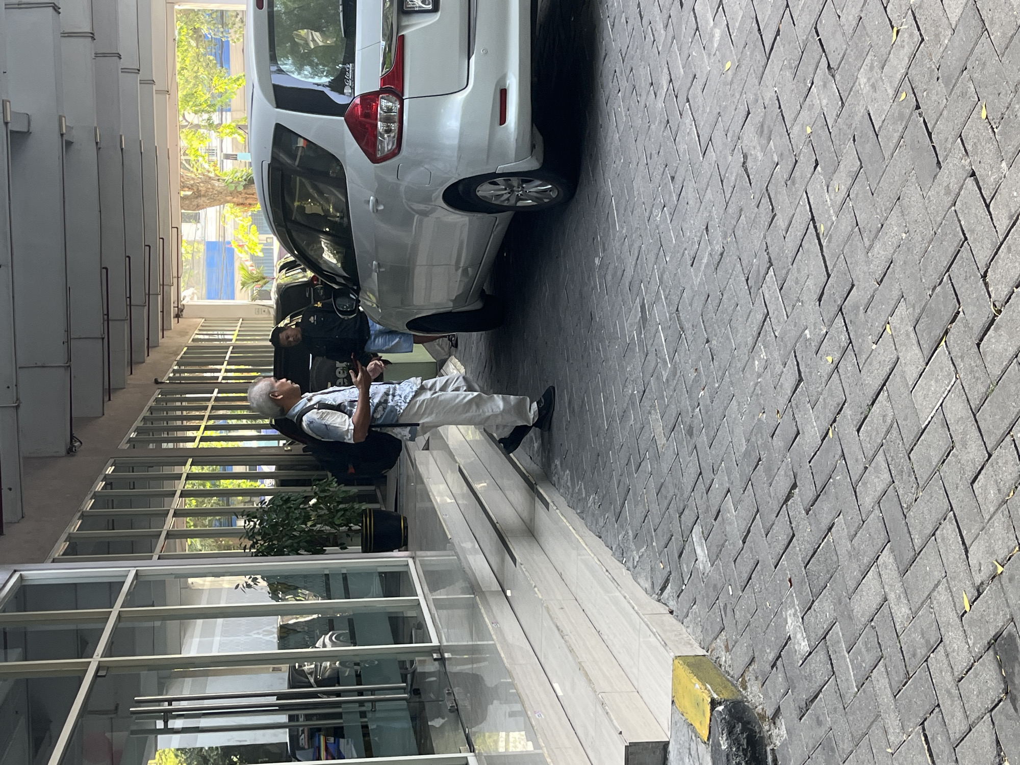 An elderly man with grey hair and a patterned shirt walks down a set of tiled stairs towards a white van parked on a herringbone-patterned brick pavement. The man carries a dark backpack over his right shoulder and wears dark shoes. To his left, a man in a black jacket and blue shorts stands near the van, holding a phone. The scene appears to be in a covered outdoor area, possibly a parking lot or entryway, with a glass-fronted building visible on the left and trees and sunlight in the background.