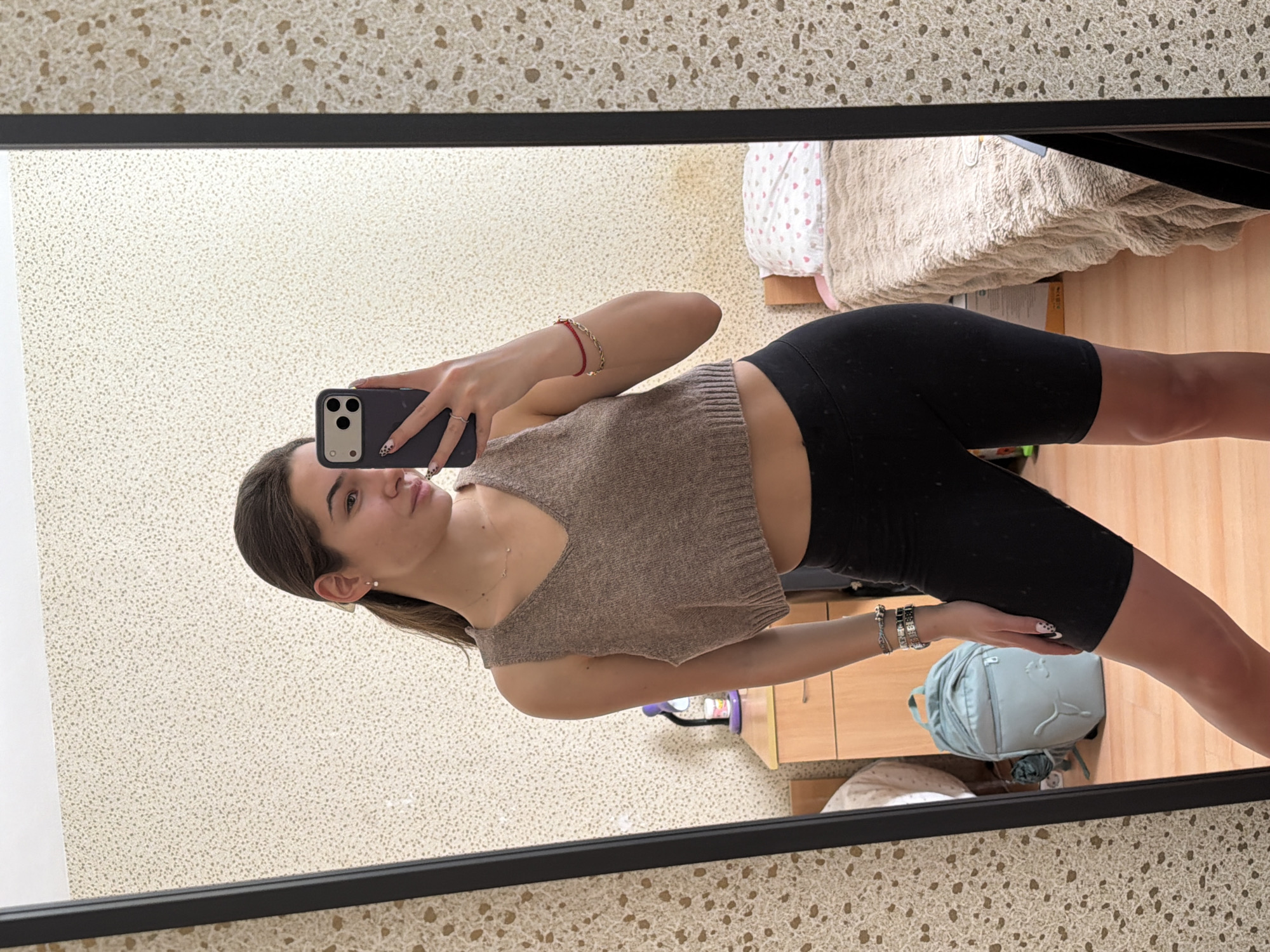 A young woman is seen in a mirror selfie in what appears to be an indoor setting. She is wearing a beige knitted top and black cycling shorts. The environment shows elements of a bedroom, with a bed and a light blue backpack visible. Based on the style of the interior, the type of electrical outlet, and the general ambiance, it is most likely the location is in Sofia, Bulgaria.