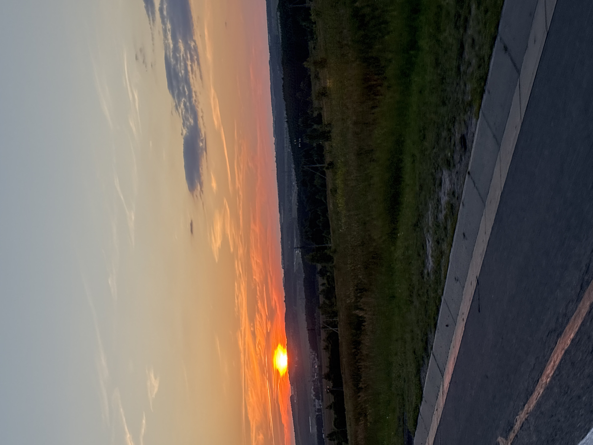 The image shows a sunset over a rural landscape, likely in or near Mayevka, Russia. The sun is a bright, intense orange, low on the horizon, casting a warm glow across the sky which transitions from orange to pale blue. Wispy clouds are scattered across the sky. The foreground features a dark asphalt road with a faded orange line, bordered by a concrete curb and grass. Beyond the road, a field of dry, golden grass stretches towards a distant line of dark trees. In the middle ground, hills are visible, dotted with small structures suggesting a village or scattered settlements. The overall impression is one of tranquility and the end of a day in a rural setting.