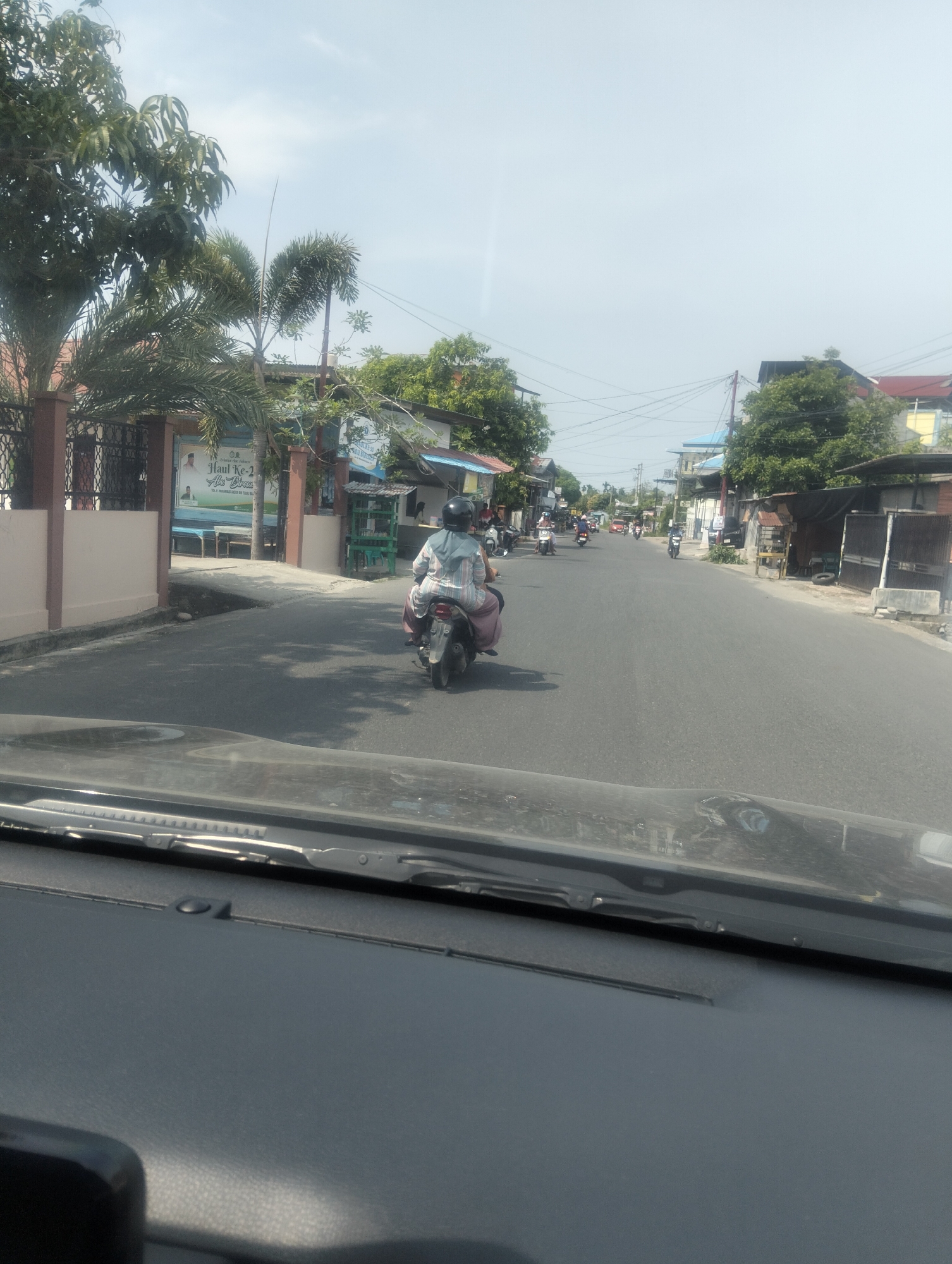This is a street-level, wide-angle shot taken from inside a vehicle, looking forward along a paved road in Geulanggang Teungoh, Indonesia. The day is bright and sunny with a clear, pale blue sky. The road is lined with buildings and vegetation.
In the foreground, the dashboard of the vehicle is visible, with a windshield wiper at the top. A black object, possibly a phone mount or part of a speaker, is in the bottom left corner.
On the left side of the road, a building with a cream-colored facade and a brown fence is visible, with a poster on the wall featuring two men and Indonesian text. Palm trees and other leafy trees are abundant.
On the road itself, a person on a silver scooter is riding away from the camera, wearing a helmet and a light-colored patterned outfit. Further down the road, several other scooters and their riders are visible, some with passengers. There are also glimpses of other buildings, including one with a blue roof and another with a dark corrugated metal roof. The road appears to be a residential or commercial street, with moderate traffic. Utility poles and wires are present along the roadside. The overall impression is a typical sunny day in a Southeast Asian urban or semi-urban environment.