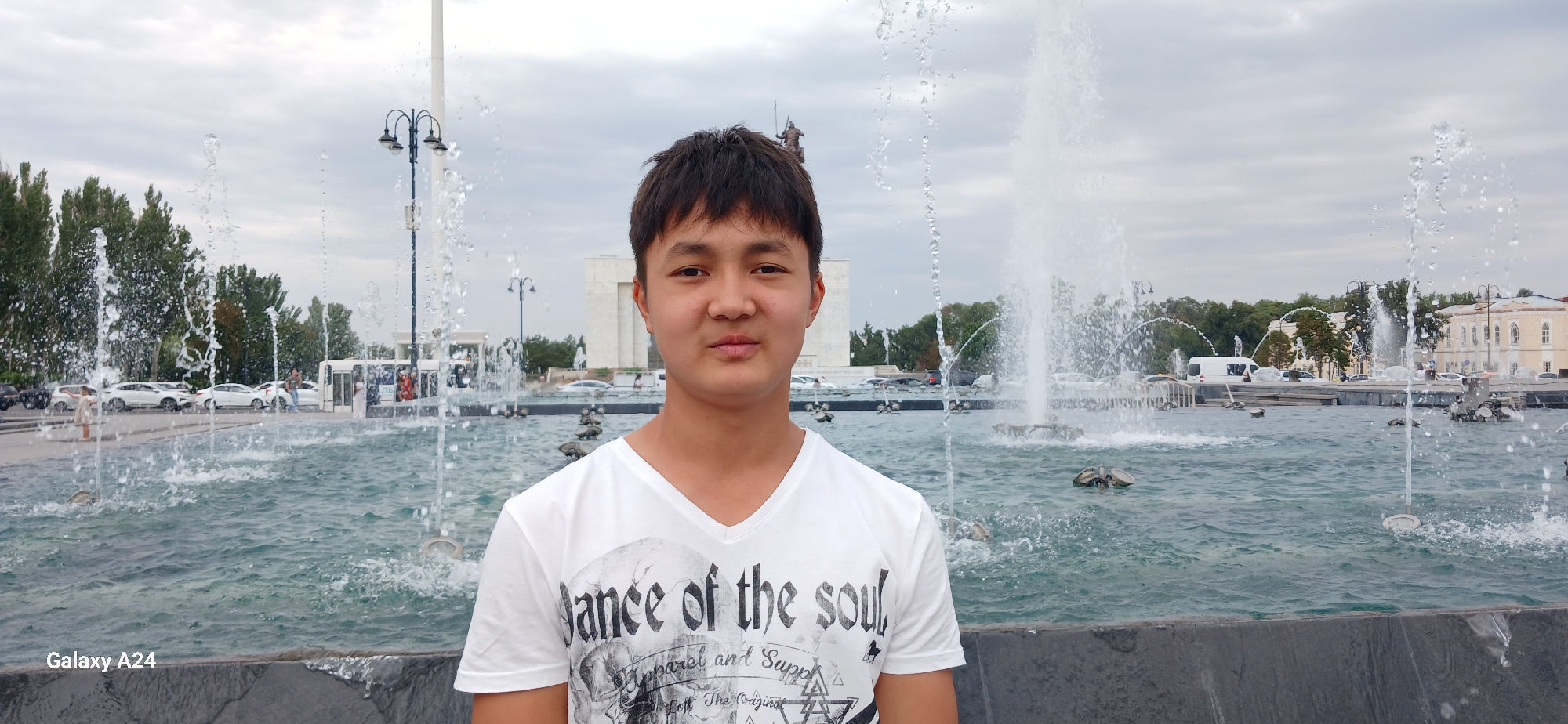 This is a waist-up, medium shot of a young East Asian male standing in front of a large fountain in Bishkek City, Kyrgyzstan. The man has short, dark hair and is wearing a white V-neck t-shirt with a graphic print that includes the words "Dance of the soul" and other decorative elements. He is looking directly at the camera with a neutral expression.
The fountain is the dominant feature of the background, with several jets of water spraying upwards. The water in the fountain is a deep blue-green color and appears choppy. Beyond the fountain, there is a street with several white cars parked and moving, a few trees, and some buildings. The sky is overcast with gray clouds. The ground in the foreground is a dark gray, textured surface. On the bottom left of the image, the text "Galaxy A24" is visible.