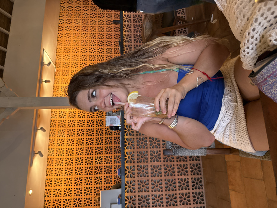 A young adult female is depicted seated in an establishment in Uluwatu, Indonesia. She has long, wavy, light brown and blonde hair and is wearing a blue strapless top and a white crocheted mini-skirt. Her left wrist displays multiple silver bracelets, and her right wrist has a silver cuff bracelet and a red string bracelet. She holds a clear, ribbed glass containing a dark liquid beverage with ice and a lemon slice, with a straw at her lips and her tongue partially extended. Her gaze is directed upwards and to her left. The background features an orange wall constructed from decorative concrete blocks with a repeating floral pattern. Above, a wooden beam supports several track lights. A bar counter, including a point-of-sale system, is visible on the left. The back of another individual, wearing jeans and a dark top, is partially visible on the right. The floor is tiled, and a woven chair back is next to the primary subject. A light-colored crocheted item rests on a wooden table in the immediate foreground.