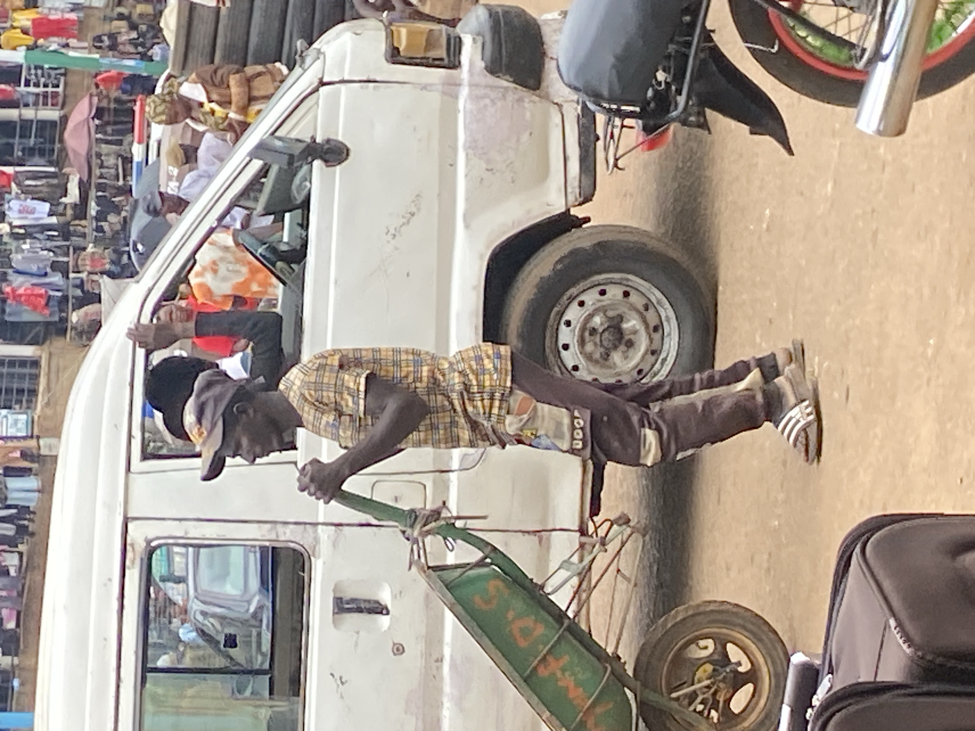 In Bukuru, Nigeria, a man in a checkered shirt and dark pants pushes a green wheelbarrow. He is positioned on the right side of the frame, facing left, with his hands on the wheelbarrow's handles. The wheelbarrow is loaded with some items and has red lettering on its side. Behind the man, a white van is visible, with its passenger door open. Several people are partially visible inside and around the van. The scene appears to be on a dirt road or marketplace, with buildings and stalls in the background. A motorcycle is also present in the lower right portion of the image.