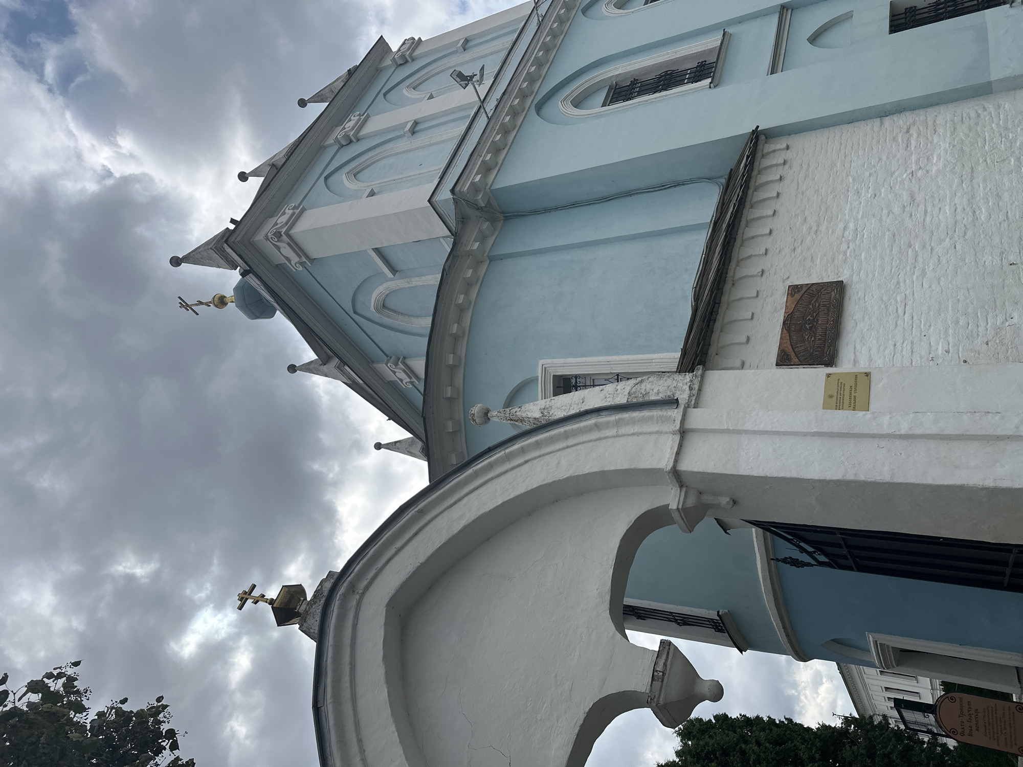 The image shows a detailed view of the blue and white building of the Novo-Golutvin Monastery in Kolomna, Russia. The architecture features arched windows, decorative white trim, and a golden cross atop a dome. The foreground includes a white arched entryway with a dark metal gate. A sign with Cyrillic text is visible on the white brick wall. The sky is overcast with grey clouds. A tree branch with green leaves is visible in the upper left corner.