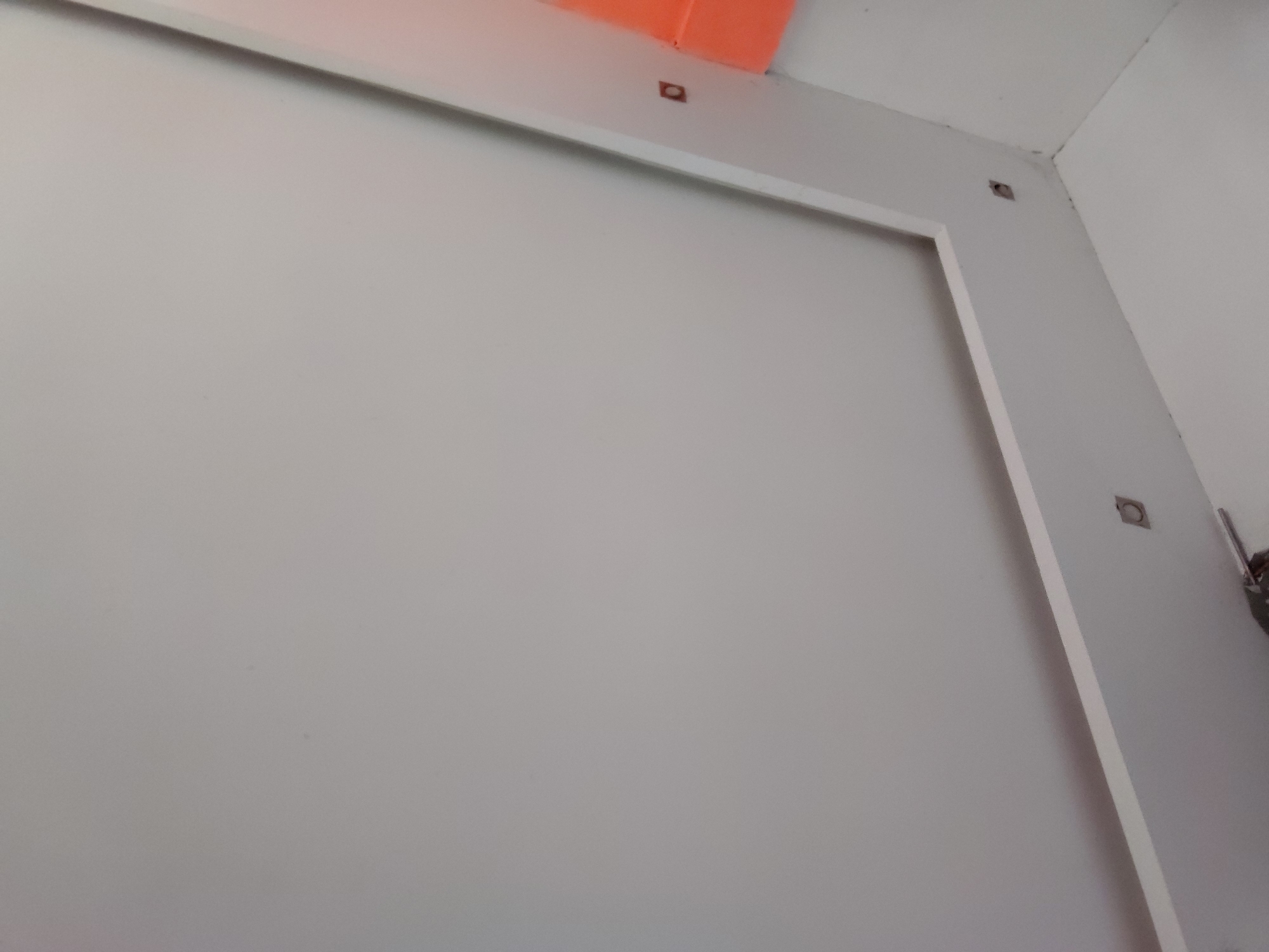 The image displays an interior wall or partition in Santa Ana, Venezuela. The central focus is a large, flat, light-colored surface, likely off-white or light grey. This surface is bordered by a clean white trim, forming a rectangular frame around a recessed or delineated panel.
Along the upper edge of the framed section and extending down the right side, several small, square-shaped fixtures or holes, dark in color, are visible. An orange-colored object is partially visible along the top edge of the frame, possibly an overhead pipe, duct, or another ceiling-mounted element.
In the upper right corner of the image, two light-colored walls converge, showing a seam with some minor dark discoloration or marks, possibly dirt or mildew. A small, indistinct dark object with a metallic reflection is also visible at the extreme right edge of the frame, suggesting a piece of equipment or fixture mounted nearby.
The lighting appears even, characteristic of an indoor setting with either artificial illumination or well-diffused natural light. No people are present, and no specific activities are depicted. There is no visible text within the image. The scene presents a static view of a functional or minimalist architectural interior.