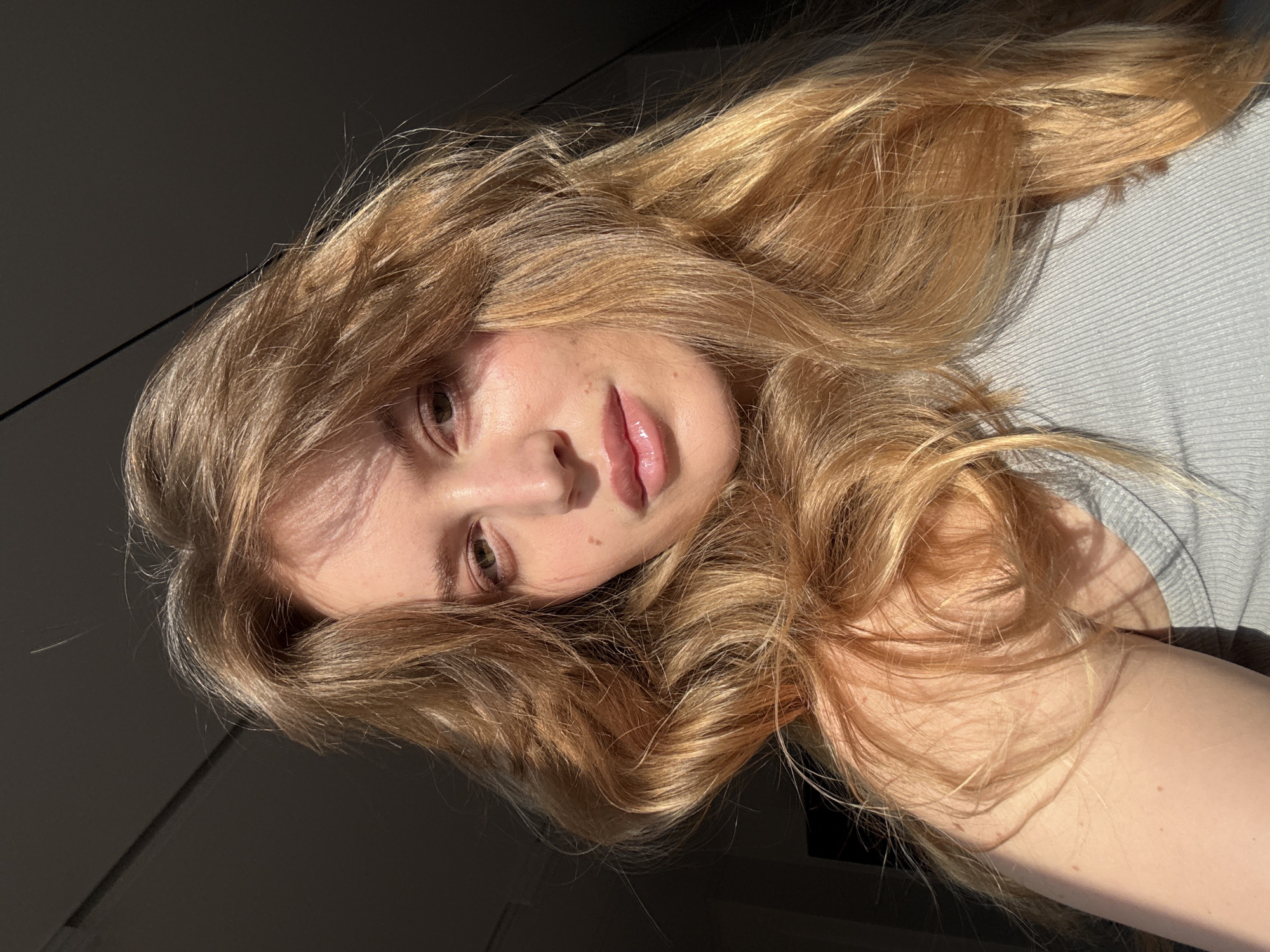 A young woman with long, blonde hair lies on a gray surface. Her face is tilted towards the camera, displaying fair skin and natural-looking lips with some shine. The left side of her face is in shadow, while the right is illuminated by direct sunlight. The background is a mix of dark and light areas, suggesting indoor lighting conditions. No location in Hrodna, Belarus is discernible from this image.