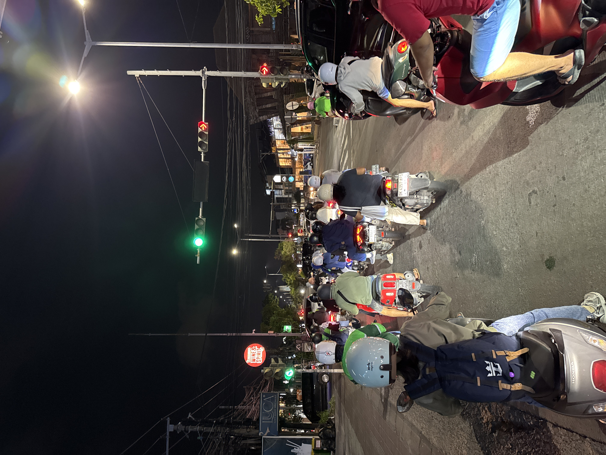 The image depicts a traffic jam at night on a street in Kuta, Indonesia. A long line of motorcycles and scooters are stopped at a traffic light. The traffic light is green for the direction facing the viewer and red with a right-turn arrow for the cross traffic. The street is paved, and there are buildings and streetlights visible on either side of the road. The sky is dark, indicating it is nighttime. Several people are on the motorcycles, wearing helmets and casual clothing. The overall scene is one of urban traffic congestion in a tropical location.