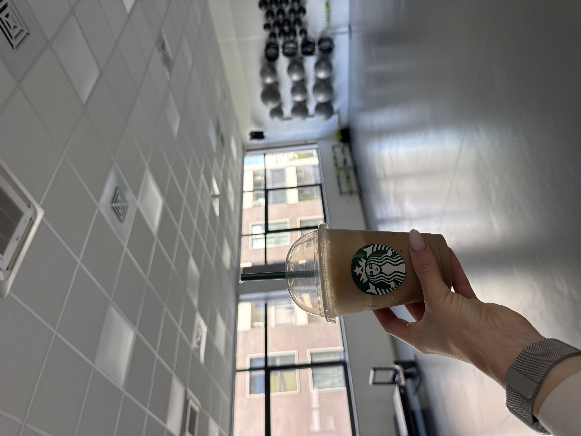 The image shows a person's hand holding a Starbucks Frappuccino in what appears to be a fitness studio or gym. The studio has a grey, reflective floor, a white ceiling with grid lighting, and large windows with a view of an apartment building. Various gym equipment, including stability balls and what looks like a weight rack, are visible in the background. The person is wearing a watch on their left wrist, and their fingernails are painted a light pink. The Starbucks logo is clearly visible on the cup. The overall lighting suggests an indoor environment with natural light coming from the windows.