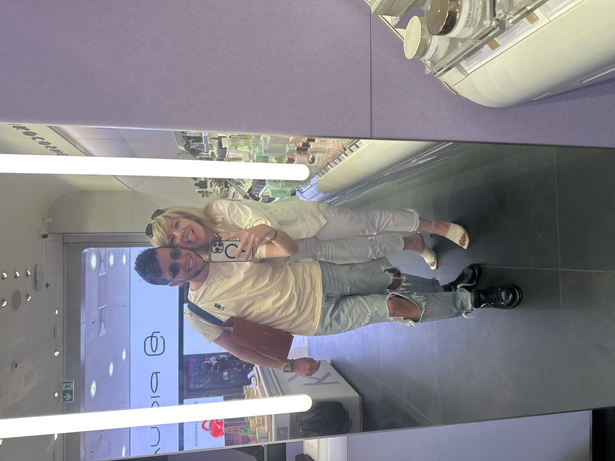 A man and a woman, a couple, take a mirror selfie in a cosmetics store. The man, in a tan t-shirt, ripped jeans, and black boots, stands on the left. The woman, in a white top and white pants, stands to his right, embracing him. Both smile at the camera, the woman sticking her tongue out. The store appears to be in Moscow, Russia, with its clean, modern aesthetic and product displays visible in the background and reflection.