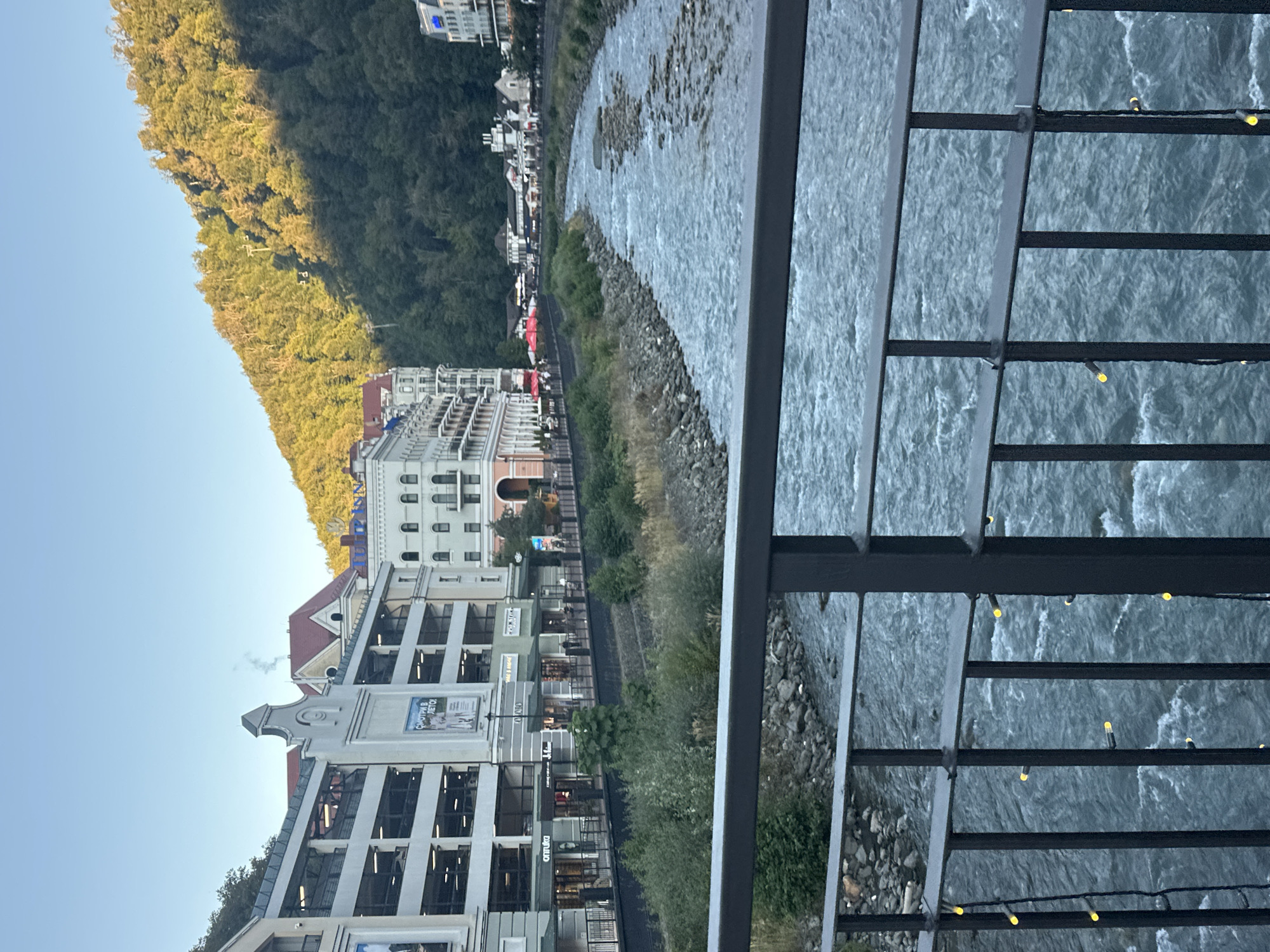 This is a wide, eye-level, exterior shot taken from a bridge railing. A river flows from the right to the left of the frame, with buildings on the left and a wooded mountain slope on the right. The river is turbulent and rocky, with the far bank lined with large rocks. The buildings on the left are multi-story, with the closest one appearing to be a parking garage, and the others being retail or residential buildings. The mountain slope is densely wooded, with trees extending to the very top of the frame, and the sun casting dappled light on the foliage. A faint wispy smoke plume rises from a roof in the middle-ground. A dark metal railing with yellow string lights adorns the bottom of the frame.