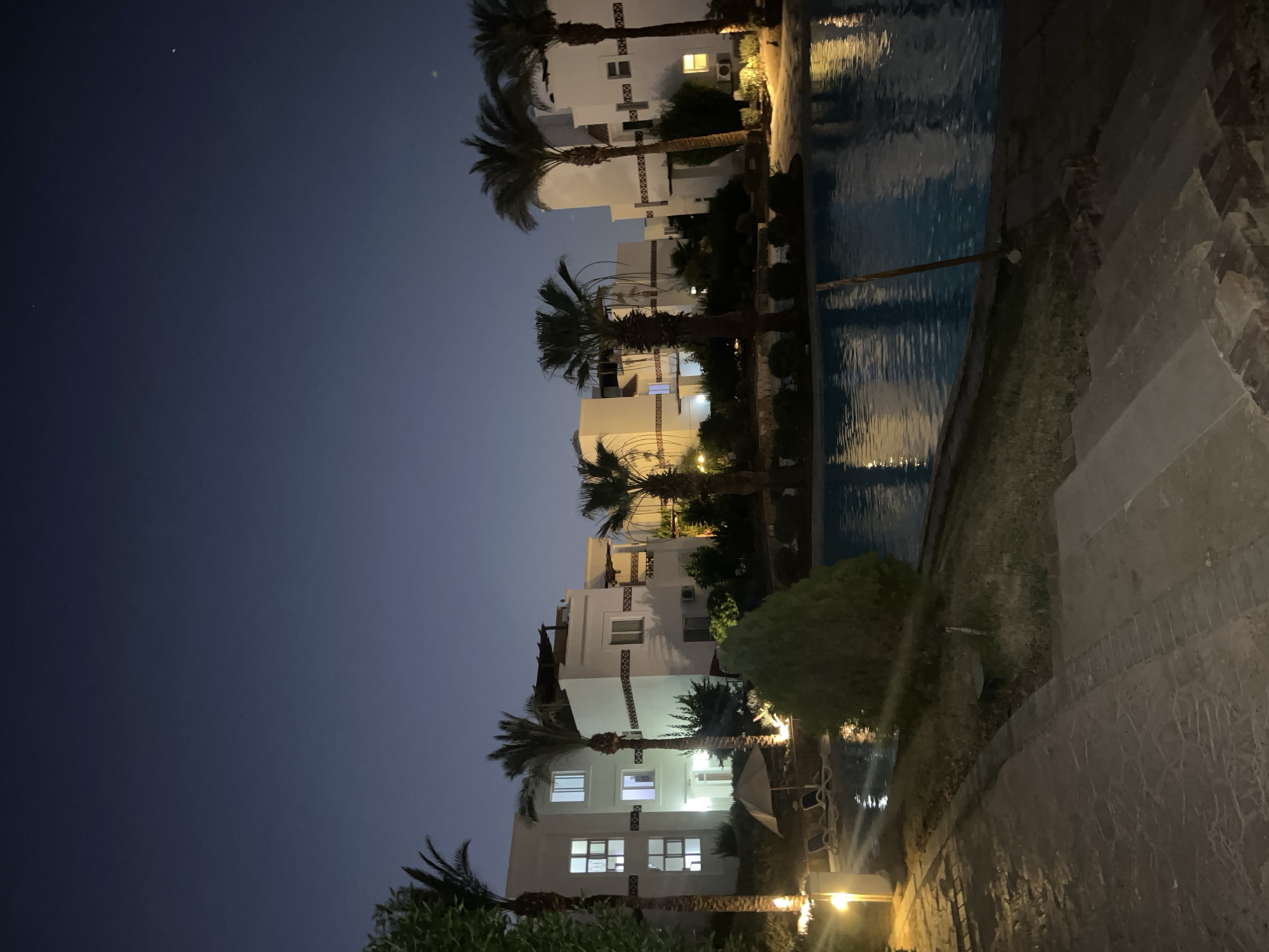 The image depicts a nighttime scene at a resort in Sharm El Sheikh, Egypt, likely overlooking a swimming pool and adjacent buildings. The sky is a deep twilight blue with a few faint stars visible. In the foreground, a paved pathway with some grass and low shrubs lines the edge of a brightly lit swimming pool. Several lounge chairs are placed near the pool, some occupied by umbrellas. Palm trees are scattered throughout the scene, their silhouettes contrasting with the illuminated white buildings in the background. These buildings appear to be multi-story accommodations with numerous windows, some of which are lit from within. The overall atmosphere is serene and inviting, suggesting a vacation resort environment.