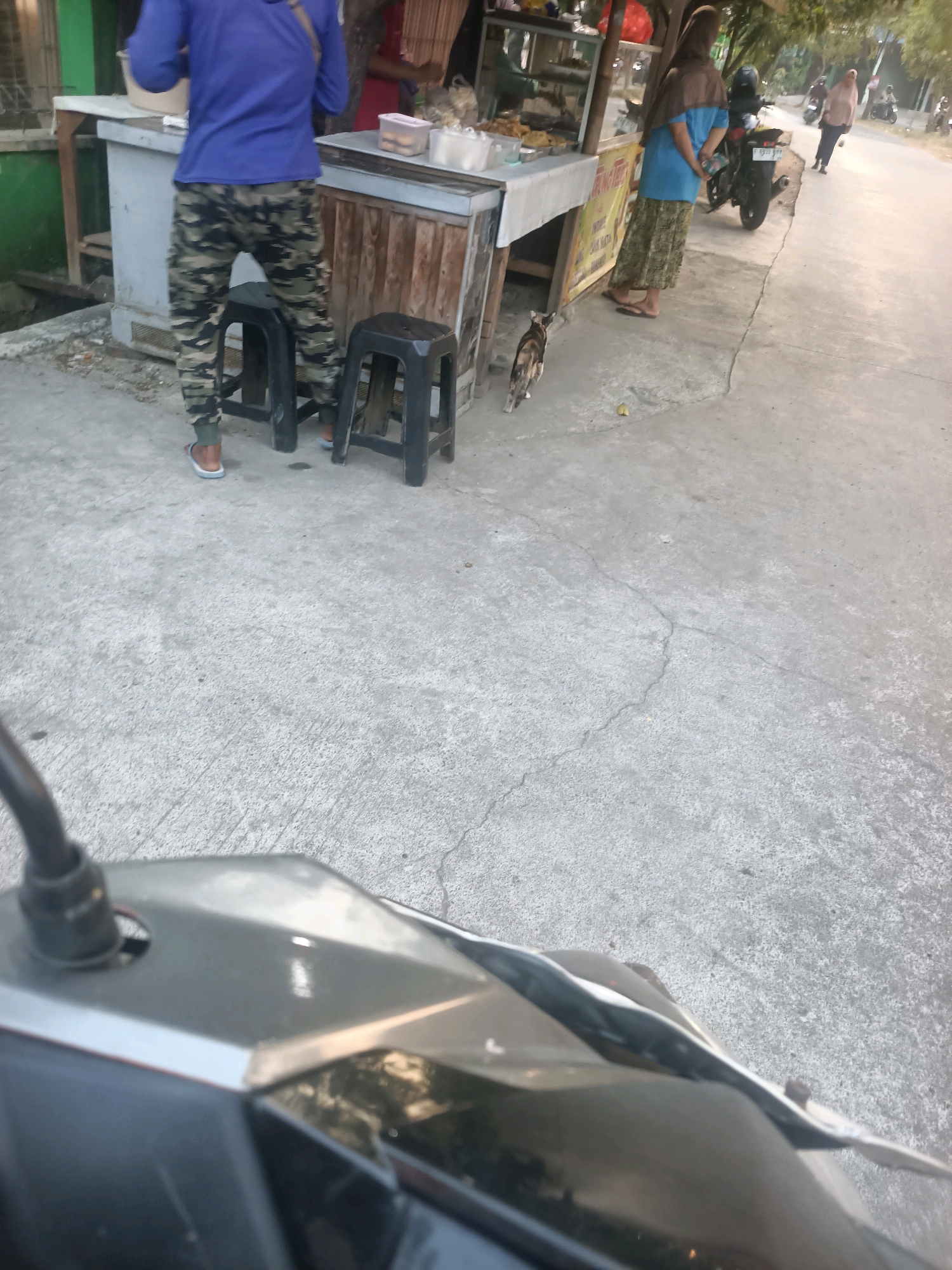 This is a street-level, point-of-view shot from a motorcycle, looking towards a street food stall in Telagaasih, Indonesia. A person in camouflage pants and a blue long-sleeved shirt stands at the stall, which has a counter displaying food items. Two black stools are placed in front of the counter. A tabby cat is walking on the pavement near the stall. In the background, two women are standing near a parked motorcycle and further down the street, several motorcycles are visible. The environment appears to be a roadside commercial area with concrete pavement and some trees. The lighting suggests it might be late afternoon or early morning.