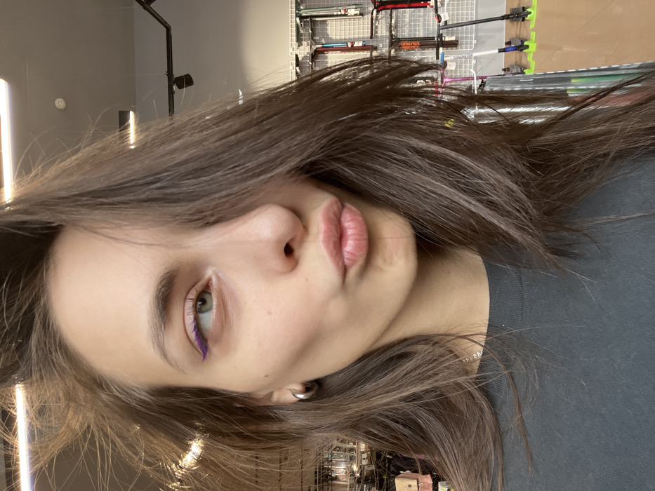 A female individual is depicted in a medium-close-up shot, gazing upwards and to the right with her left eye, which features a prominent purple winged eyeliner. Her lips are puckered into a "kissing" pose. She has long, brown hair, partially obscuring the right side of her face, with some strands appearing disheveled on top. A silver-colored hoop earring is visible on her left earlobe. She wears a dark, possibly charcoal, crew-neck t-shirt and a thin silver-colored necklace. The background reveals a retail store interior with modern strip lighting and track lights on a light-colored ceiling. To the right, a white grid-panel display wall showcases various scooter components and complete scooters. The setting is identified as a Samokat shop (scooter store), likely at Prospekt Svobodny, 48A, Krasnoyarsk, Russia.
