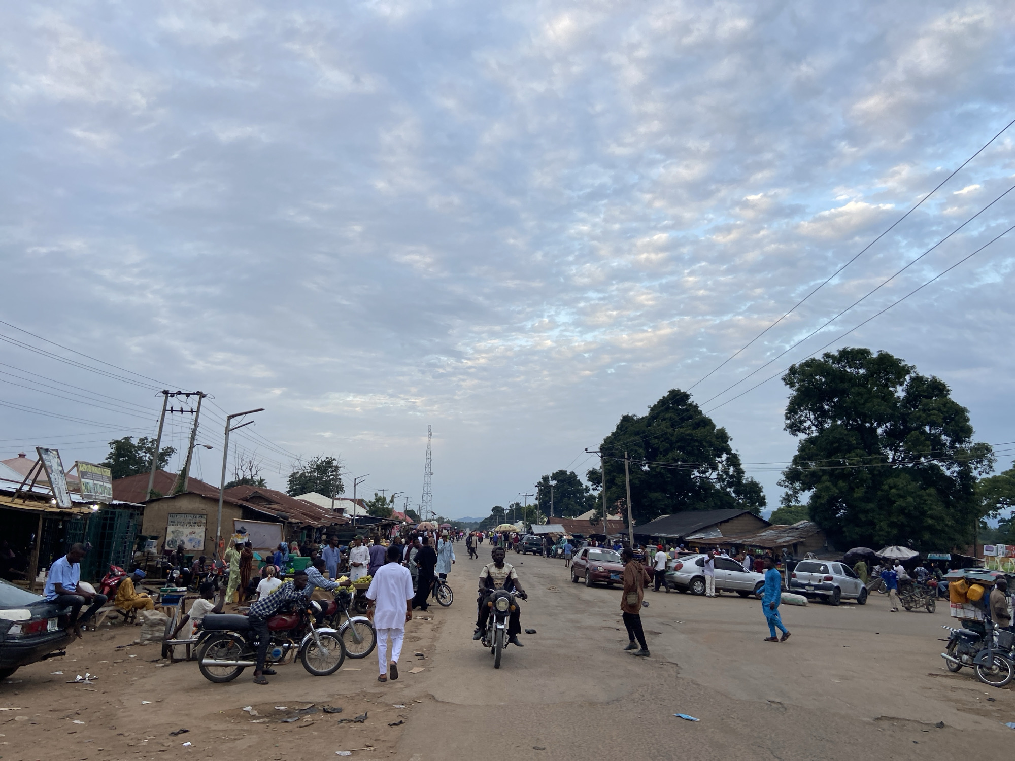 The image depicts a bustling street scene in Mararaba, Nigeria. The sky is overcast with a textured cloud cover. The street is unpaved, with visible dirt and debris. Several people are walking, riding motorcycles, or congregating on the sides of the road. A man in white clothing is walking towards the camera. A man is riding a motorcycle in the middle of the road. There are several parked cars along the side of the street. Buildings line the street, and some appear to be makeshift or simple structures. Trees are visible in the background. Power lines stretch across the sky. A tall telecommunications tower is visible in the distance.