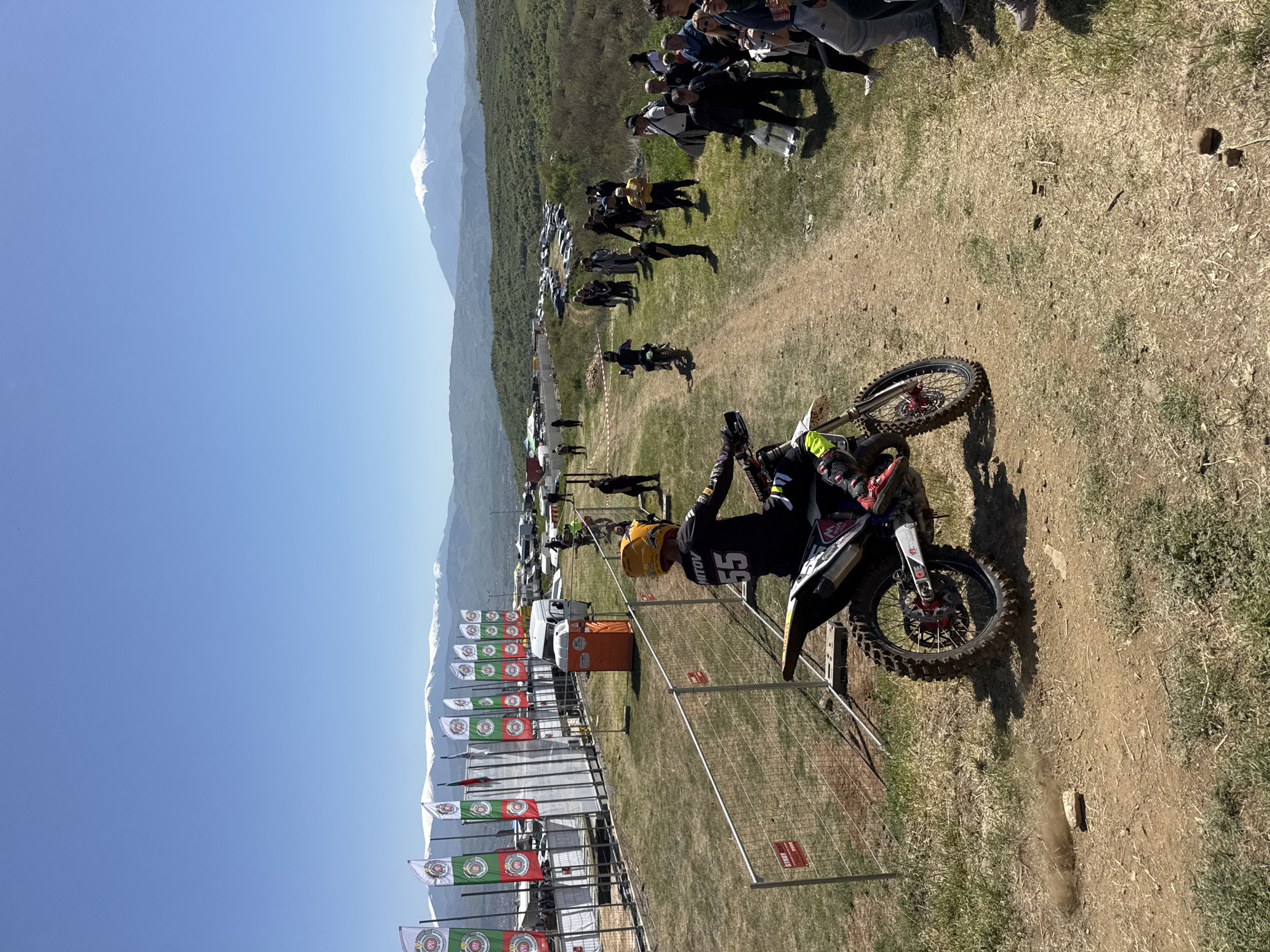 The image shows a motocross event taking place in Selishte, Bulgaria. A motorcyclist wearing a black racing suit and a yellow helmet, with the number 55 visible on the back, is riding a dirt bike. The event is marked with flags displaying a logo and fencing. Spectators are visible on a grassy hill beside the track. In the background, there are mountains and a clear blue sky. There are vehicles parked behind the event area.