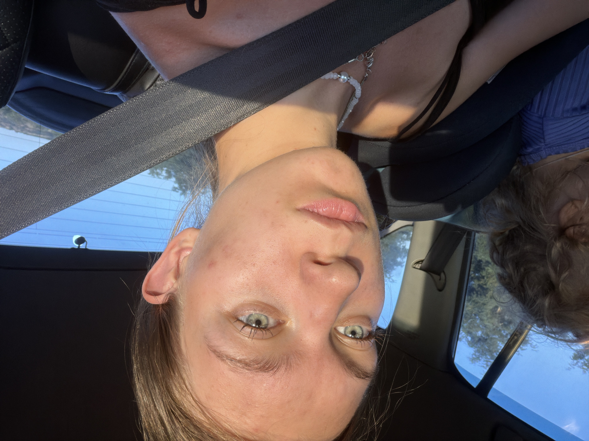 A young woman with blonde hair and green eyes is in the passenger seat of a car. She is looking at the camera, and her face is illuminated by sunlight. She is wearing a black tank top and a beaded necklace. A seatbelt crosses her chest. In the background, the sky is blue, and trees can be seen through the car window. Another person's shoulder and hair are visible in the left side of the frame. The location of Belogorki, Russia is not depicted in the image.