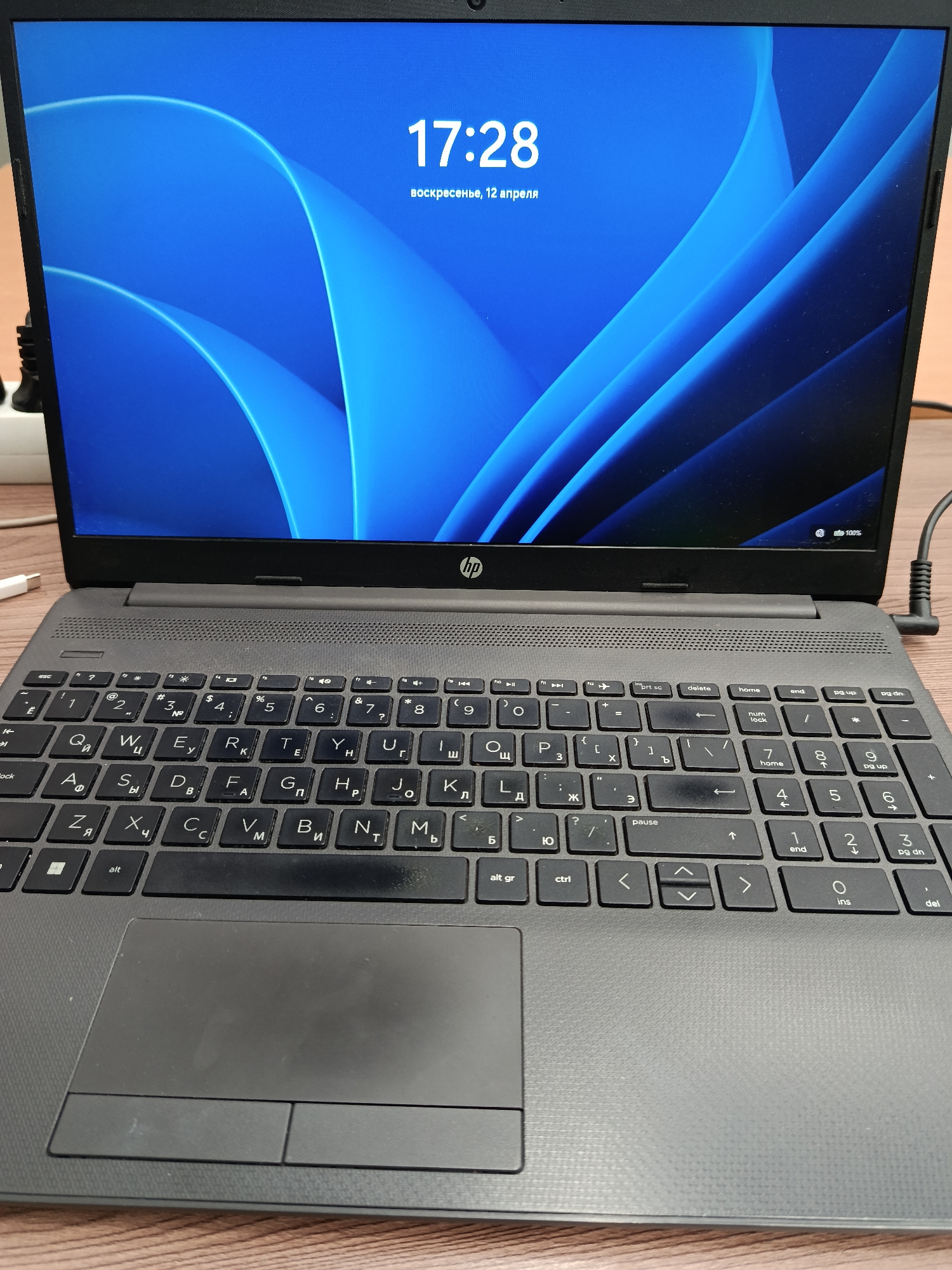 Here's a detailed description of the image:
**Overall Impression:** The image depicts an HP laptop computer resting on a wooden surface. The screen is on, displaying a blue abstract background with the time "17:28" and the date "воскресенье, 12 апреля" (Sunday, April 12th).
**Objects:**
* **Laptop:** An HP laptop is the central object. The keyboard layout is in Cyrillic, indicating it's likely intended for a Russian-speaking user.
* **Screen Display:** The screen shows a lock screen or desktop with a blue abstract pattern, time, and date. The battery level is also indicated as 100%.
* **Keyboard:** The keyboard is predominantly black with Cyrillic characters.
* **Touchpad:** A grey touchpad is located below the keyboard.
* **Power Cable:** The power cord is visible connected to the laptop.
* **Surface:** The laptop rests on a light-brown wooden surface.
* **White Cable/Adapter:** A small white cable or adapter is seen in the background, slightly out of focus.
**Environment:**
* The background is slightly blurred, indicating a shallow depth of field. The environment suggests an indoor setting, likely a home or office. The lighting is even, casting minimal shadows.
**Language:** The screen text and keyboard characters indicate the Russian language.
Based on the Cyrillic keyboard and the Russian date displayed, the laptop is most likely being used in a location where the Russian language is prevalent. Given that Moscow is the most populous city in Russia, it is a reasonable assumption that this photo was taken in Moscow, Russia.