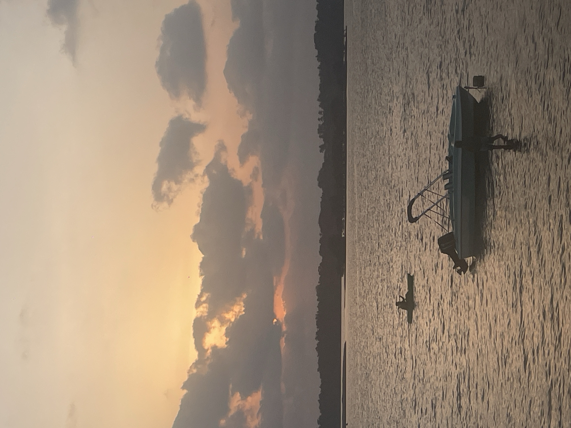 This image depicts a water scene, likely captured near Chapora, India, at dusk or dawn. The sky is filled with clouds illuminated by a muted yellow light, possibly from the rising or setting sun. On the right, a body of water extends into the scene, with visible ripples on its surface. There are two watercrafts present: a larger boat with a motor attached is positioned near the top of the frame, and a smaller boat with a person in it is farther away. The horizon line is partially obscured by a darker landmass or dense vegetation.