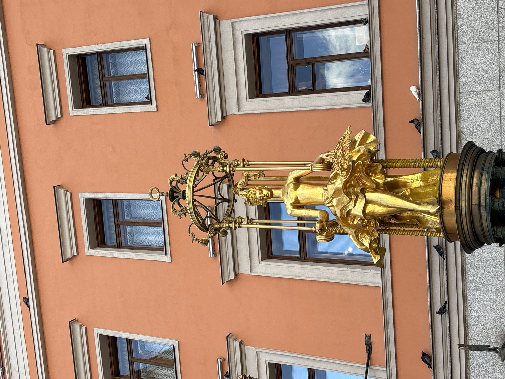 The image shows a golden statue of a woman holding a basket, situated in front of a building with an orange facade. The statue is elaborate, with the woman wearing a flowing dress and adorned with a crown. The building features several windows with white trim and dark wooden frames. There are numerous pigeons perched on the ledges and surfaces of the building, as well as on the statue itself. The statue appears to be a fountain, with a base that suggests water features. This is the Monument to the Founders of Moscow, located on the Moskvoretskaya Embankment in Moscow, Russia.