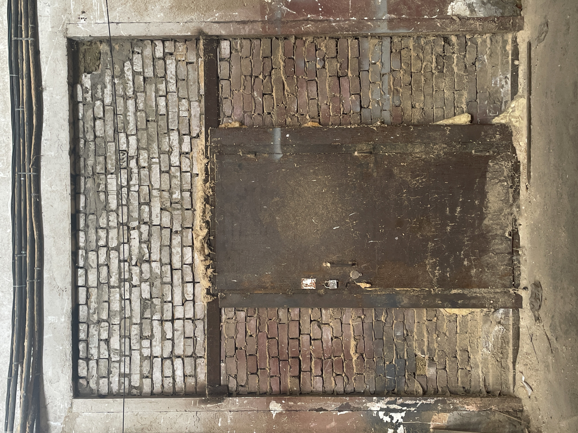 This is a top-down, close-up shot of a rough, unfinished wall surface, likely in a construction or renovation setting. The wall is predominantly composed of brickwork, with some sections appearing older and more worn than others. To the left, a section of wall is made of lighter-colored, rough-hewn bricks, partially covered in grey plaster or cement. Adjacent to this is a large, dark, rectangular object, possibly a wooden panel or metal plate, set into the brickwork. Below this central panel, more brickwork is visible, this time with darker, red-brown bricks that also show signs of age and wear, with visible mortar and some debris. To the far left of the image, a bundle of black electrical cables is neatly strapped to a light-colored vertical surface, which appears to be a concrete pillar or wall. The overall impression is one of an interior space undergoing construction or deconstruction, with exposed building materials. There is no indication of time of day or weather. No text is visible.