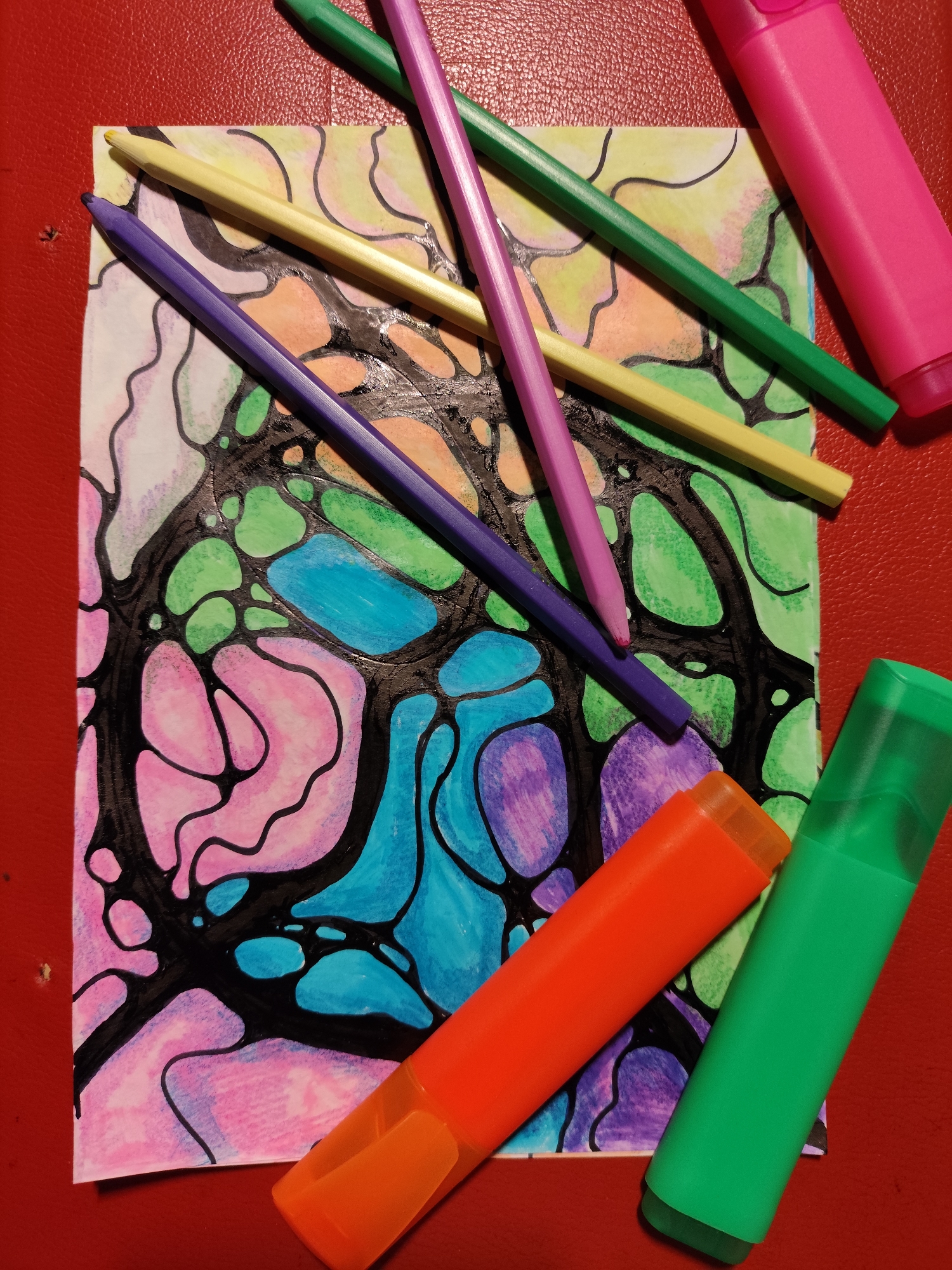The image shows a piece of art on a red textured surface. The artwork is a drawing with black outlines and filled with various colors, including pink, purple, blue, green, and yellow. The drawing appears to be a neurographic art style, characterized by organic shapes and flowing lines. Scattered around the artwork are art supplies: several colored pencils (purple, yellow, green) and two highlighters (orange and green). A pink highlighter is also visible in the upper right corner. The red surface appears to be a table or mat. There are no people or animals in the image, and no indication of a specific location in Krasnodar, Russia.