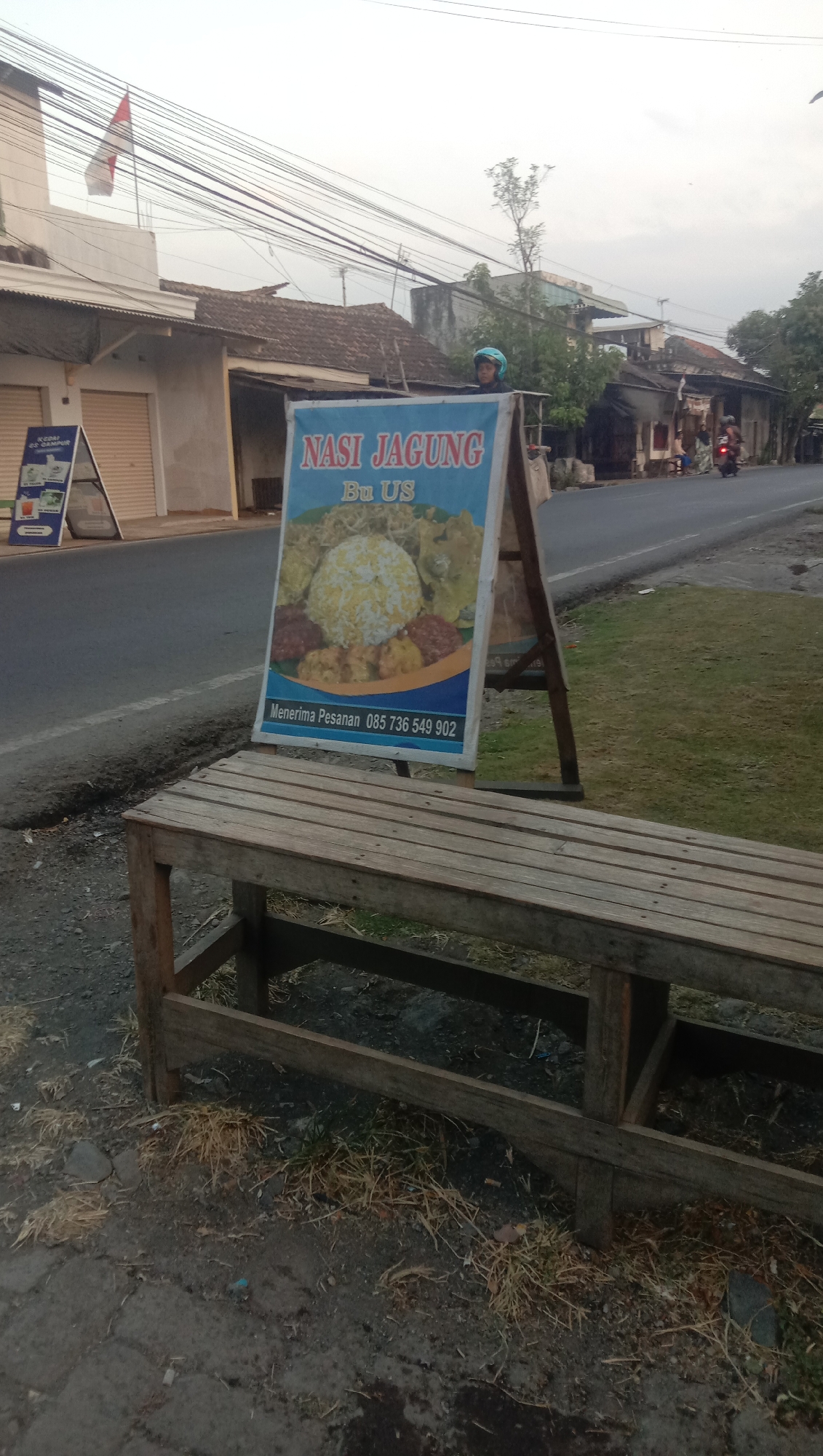 The image depicts a street scene in Karang Anyar, Indonesia. In the foreground, a weathered wooden bench sits on a gravel and paved surface. Behind the bench, an A-frame sign displays food items, with the text "Nasi Jagung Bu Us" prominently featured. The sign also includes an image of rice and side dishes and contact information "Menerima Pesanan 085 736 549 902". To the left of this sign, another A-frame sign shows a menu with drink and food options. A person wearing a blue helmet is visible behind the "Nasi Jagung" sign. The street is lined with buildings, some with closed shutters, and power lines crisscross overhead. Further down the street, a motorcycle with a rider is seen. The sky is overcast, suggesting early morning or late afternoon.