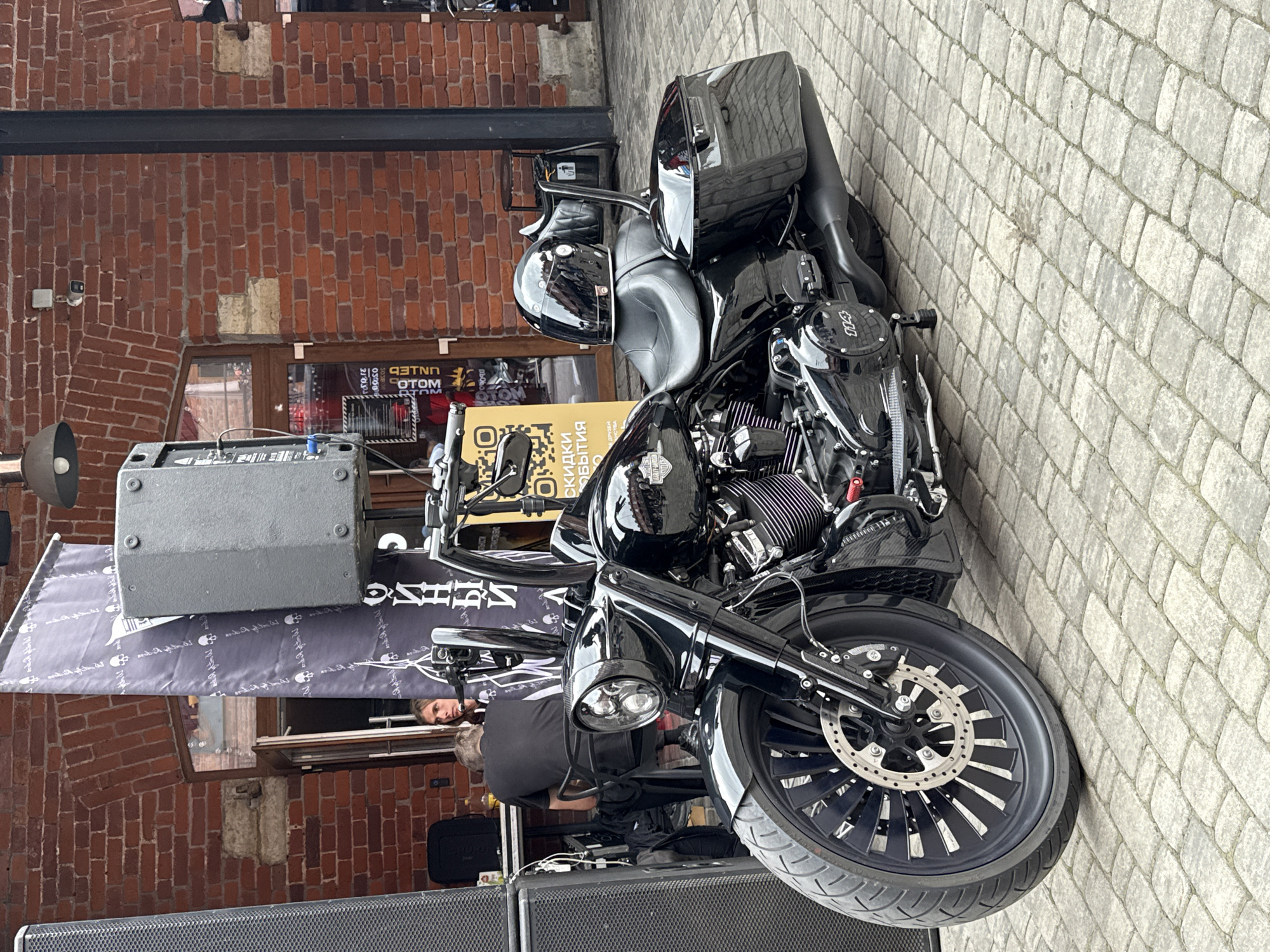 A black Harley-Davidson motorcycle is parked on a cobblestone street in front of a red brick building. The motorcycle has a black helmet on the seat and saddlebags. Two people are visible in the background, one sitting at a table and the other standing. A large speaker system is to the left of the motorcycle. The building appears to be a commercial establishment, possibly a bar or restaurant, with a banner displaying text in Russian. The scene is likely outdoors, with natural light and a brick facade typical of historic areas in Saint Petersburg, Russia.