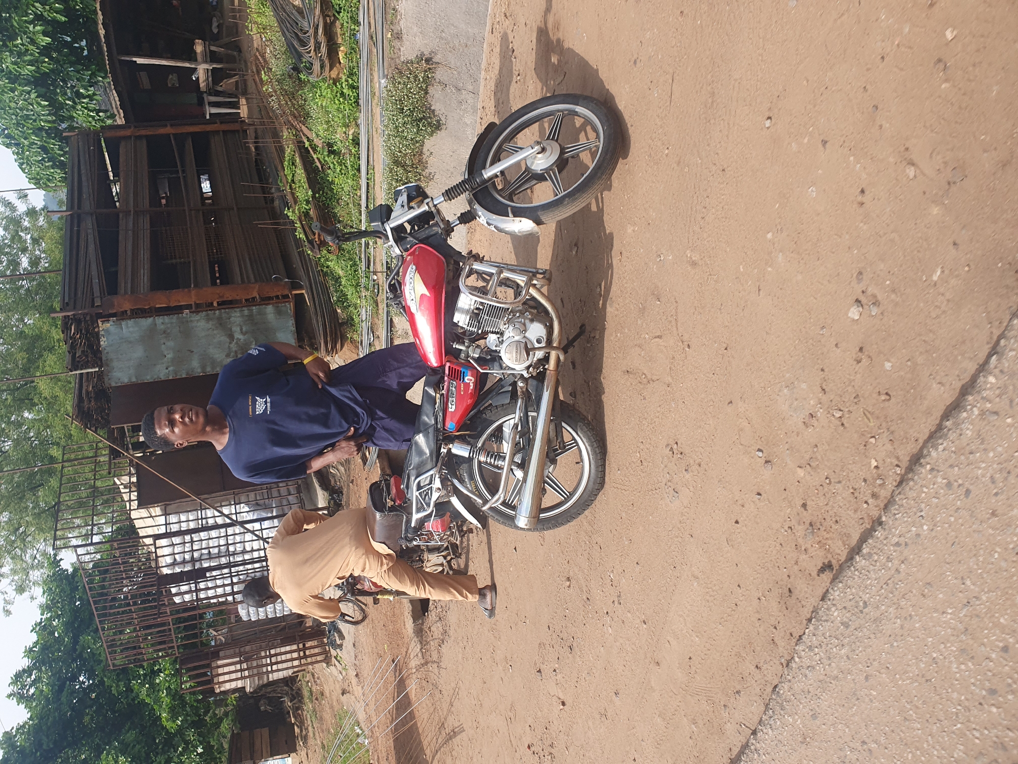 In Lokoja, Nigeria, a man in a blue t-shirt stands next to a red motorcycle. His hands are in his pockets, and he appears to be looking off to the side. Another man, dressed in a tan outfit, sits on a second motorcycle behind the first. The motorcycles are parked on a dirt road. In the background, there is a structure with metal shelving filled with long, thin objects, likely construction materials. Trees and vegetation surround the area. The scene appears to be an outdoor marketplace or a storage area for building supplies.