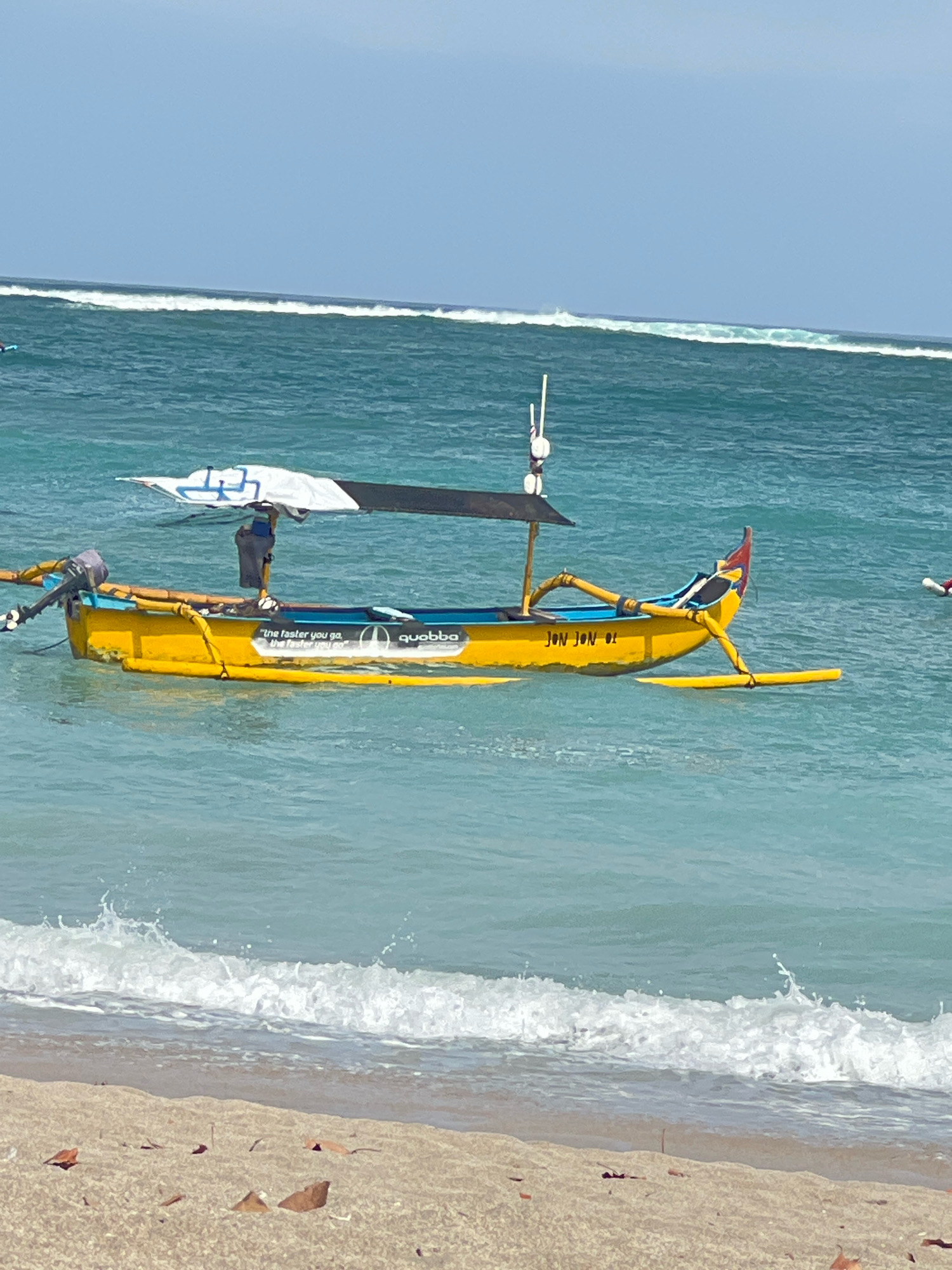 A yellow motorized outrigger canoe is floating on the turquoise waters of Kuta Beach, Bali, Indonesia, with waves breaking in the foreground and a clear blue sky overhead. The canoe has a blue interior, a black and white canopy, and an outboard motor attached to the stern. There is writing on the side of the canoe, including "quobba" and what appears to be a slogan. The name "JON JON 01" is also visible on the side. The beach in the foreground is sandy and strewn with some dried leaves and debris.