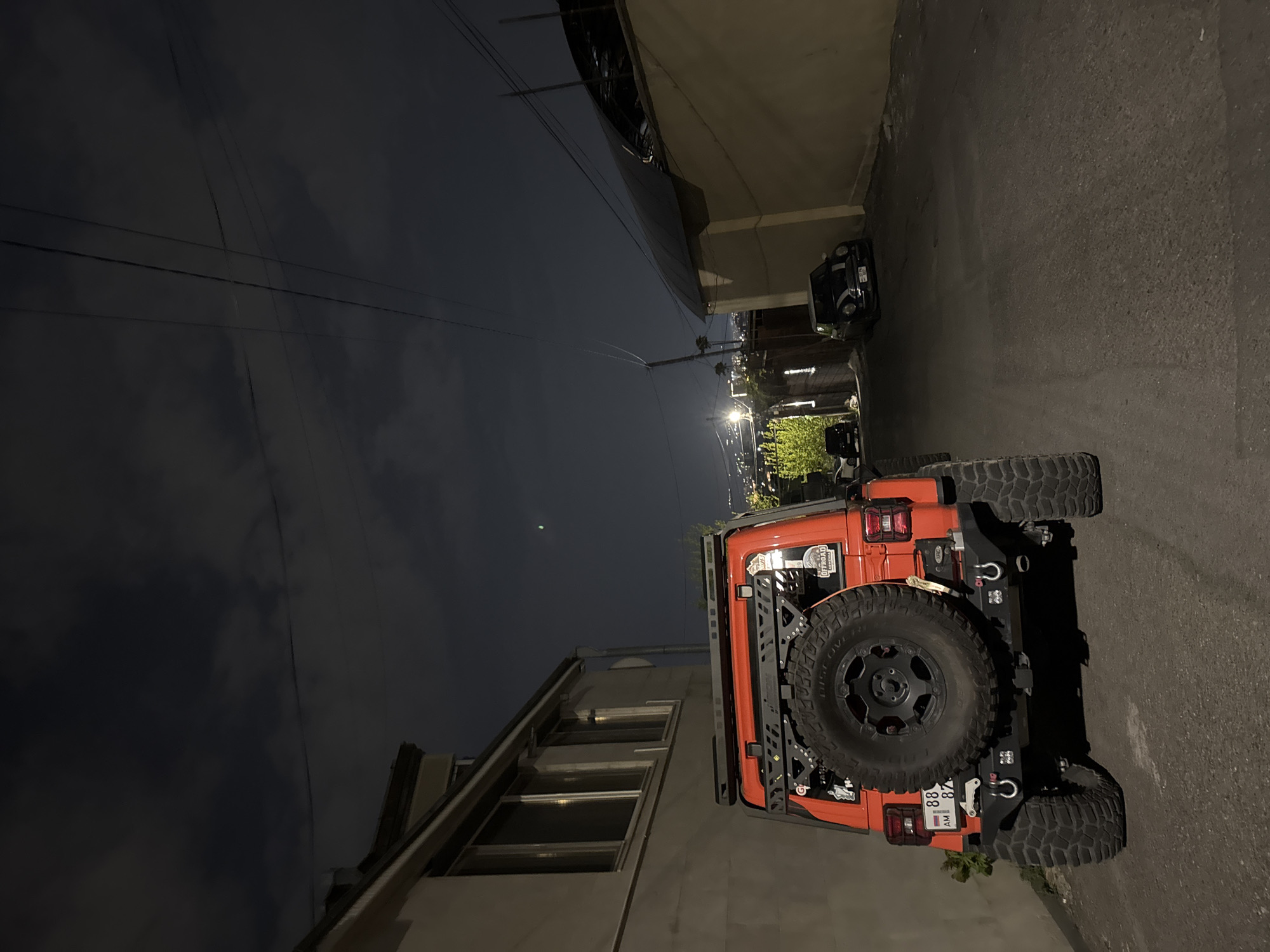 The image captures a nighttime scene on a narrow paved street in Yerevan, Armenia. An orange Jeep Wrangler is prominently parked on the right side of the street, facing away from the camera, with its rear in the foreground. The Jeep is heavily customized for off-road use, featuring large, knobby tires, a spare tire mounted on the tailgate, and a robust aftermarket rear bumper equipped with two red recovery shackles. A black ladder or side rack is visible along the left side of the vehicle. A sticker on the rear of the Jeep partially reads "OFFROAD ADVENTURES."
The vehicle's license plate, "88 AM 8847," is clearly visible. In the distance, streetlights cast a warm glow, illuminating other parked vehicles, including a smaller, dark-colored car (possibly a Mini Cooper) further down the road on the left. Buildings line both sides of the street, with a multi-story structure featuring multiple windows visible on the left. Overhead, utility lines traverse the dark night sky, which appears clear. No people are visible, and the scene conveys a quiet, static moment in the late evening or early morning hours.