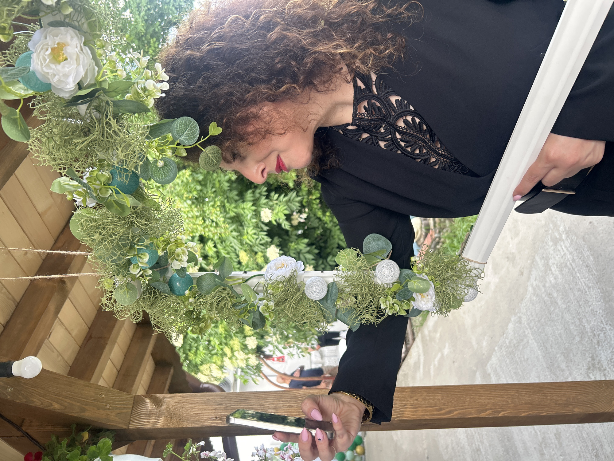 The image appears to be taken outdoors in Sandrovo, Bulgaria. A woman with curly brown hair and wearing a black top with lace detailing at the neckline is shown. She has pink nail polish. She is holding a smartphone in her right hand, which is positioned near a wooden structure. The woman appears to be inspecting or adjusting a floral wreath hanging from the wooden structure. The wreath is decorated with white and green foliage, moss, and other floral elements. The background includes greenery and possibly parts of a building or other structure. A white pole or support structure is also visible.