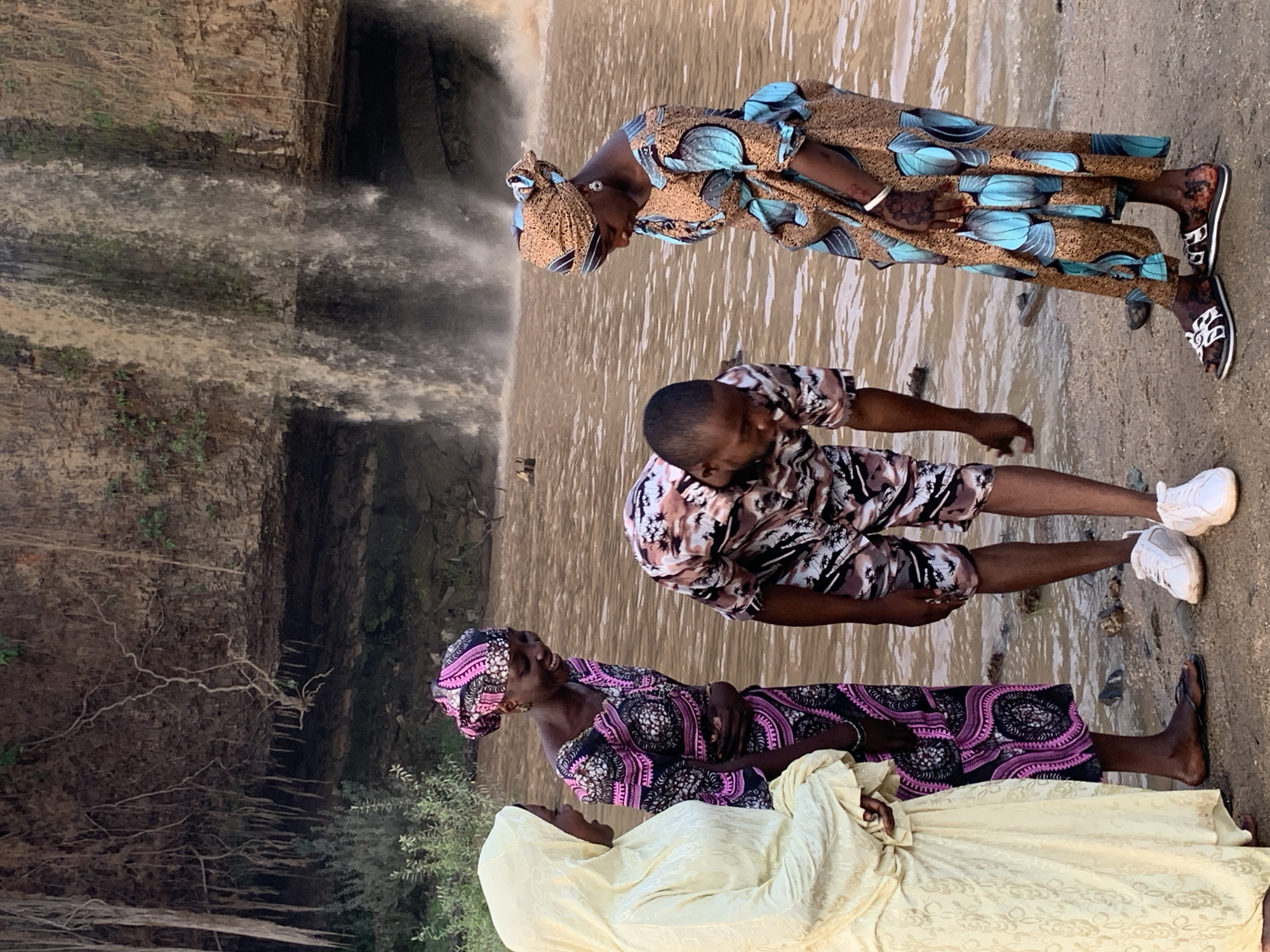 Four individuals stand near a waterfall at Gurara Waterfall in the city of Kukum, Nigeria. One woman is dressed in a blue and brown patterned robe and headwrap. Another woman wears a black and pink patterned robe and headwrap. A third woman wears a long, pale yellow robe with a hood. A man stands between the two women, wearing a tan, brown, and black patterned shirt and shorts. The waterfall is on the left of the frame. The ground is covered in mud and debris.