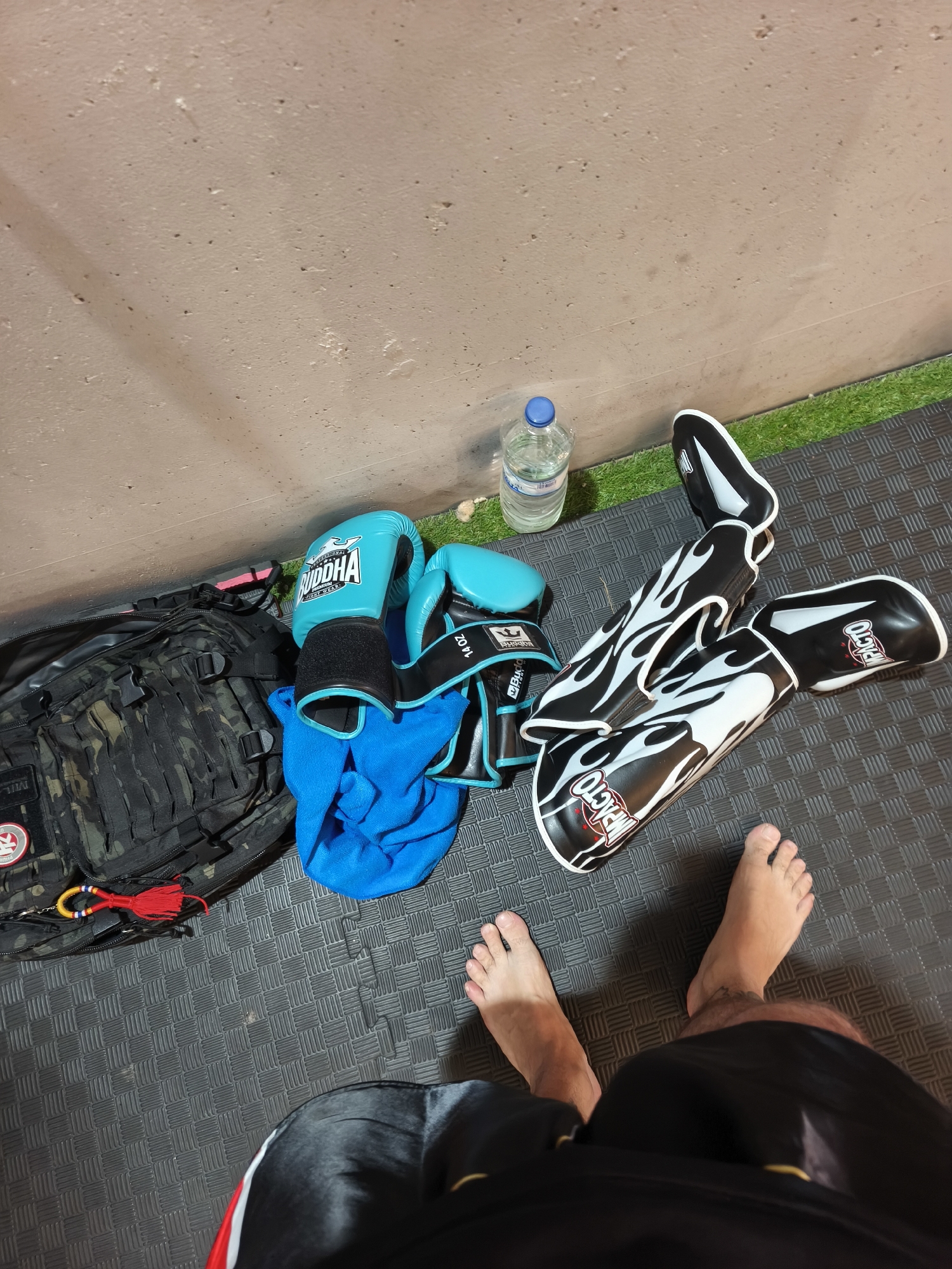 The image shows a person standing on a black interlocking foam mat in Polígon Industrial De Picassent, Spain. The person is barefoot, wearing black and red shorts. To the left of the person, there is a camouflage backpack, a pair of light blue "BUDDHA" boxing gloves, and a blue towel. Near the gloves is a bottle of water. To the right of the gloves, there is a pair of black and white shin guards that say "IMPACTO." The background includes a beige wall and a strip of artificial green grass at the bottom of the wall.