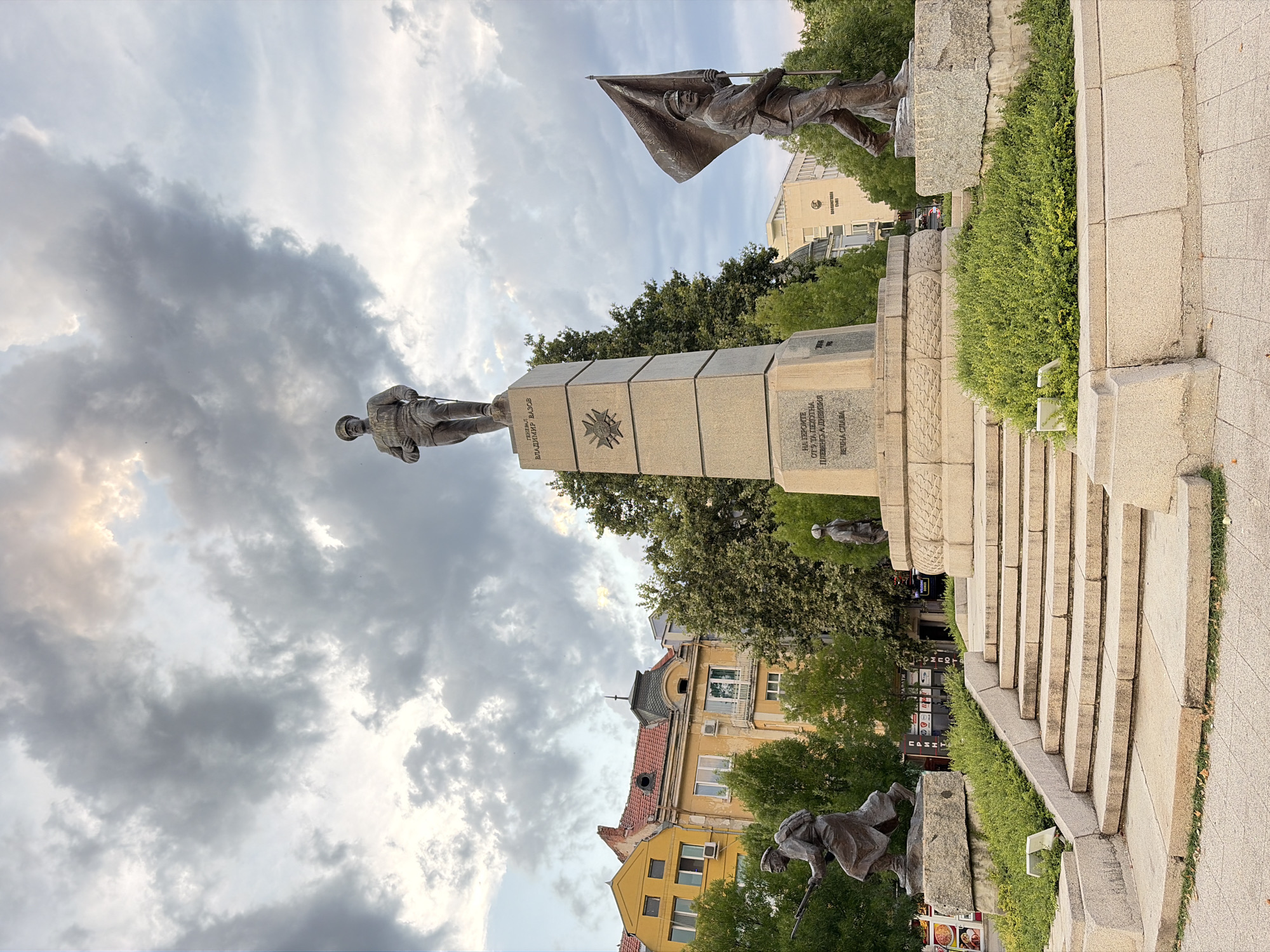 The image depicts the Monument to the Freedom Fighters in Pleven, Bulgaria. The monument features a central obelisk topped with a statue of a standing soldier in military uniform, identified by text on the obelisk as General Vladimir Vazov. Flanking the central monument are two bronze statues of soldiers. On the left, a soldier is depicted in action, carrying a rifle and a backpack. On the right, another soldier is shown marching, holding a flag aloft. The monument is set on a tiered stone base with steps leading up to it, surrounded by lush green bushes and trees. In the background, several buildings are visible, including a distinctive yellow building with a red tiled roof and a modern white building. The sky is overcast with dramatic, cloudy formations, suggesting either dawn or dusk. The scene is static, with no people actively interacting, and the focus is on the historical monument and its setting.