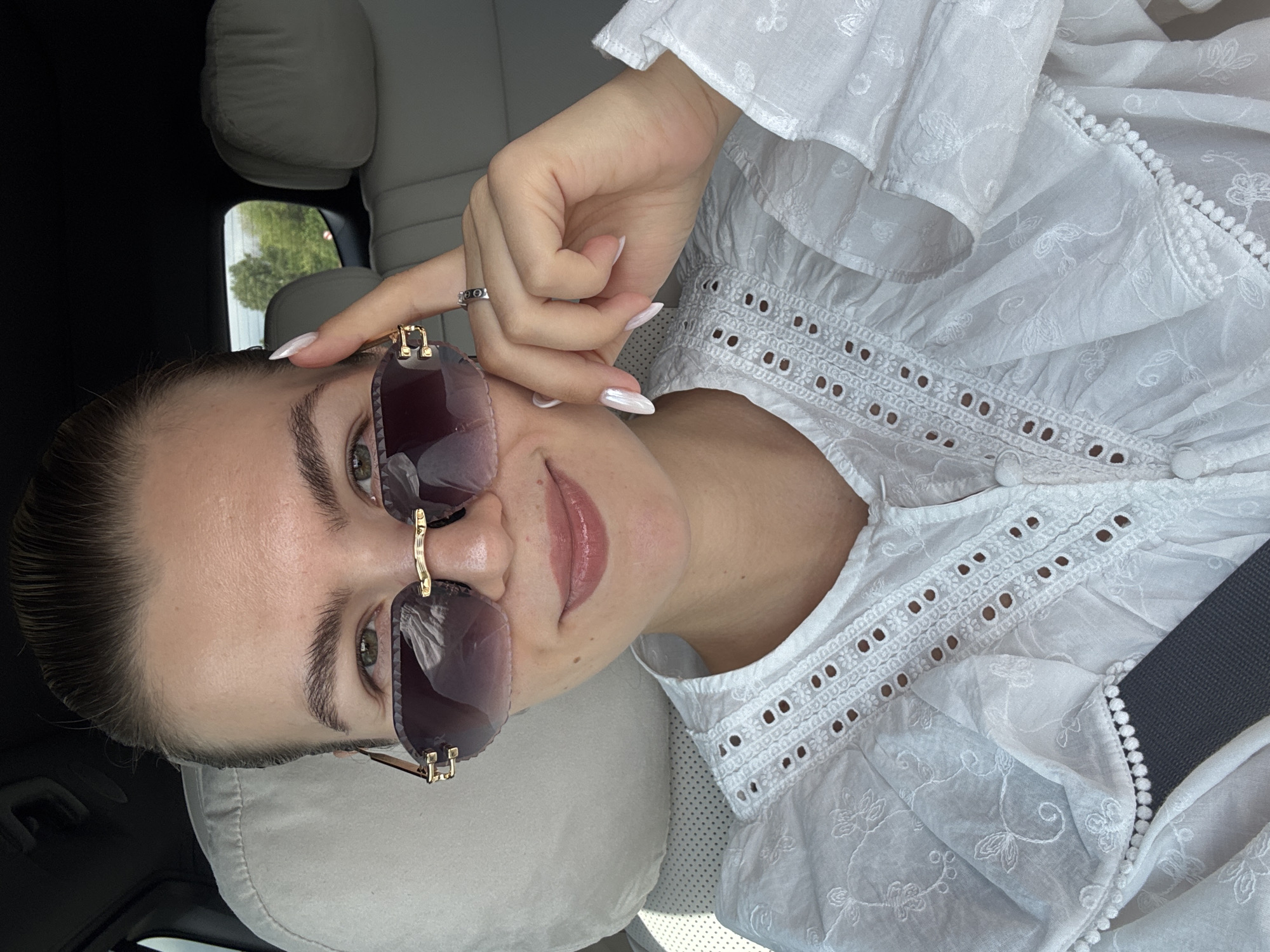 A woman with her hair pulled back is sitting in the passenger seat of a car, wearing sunglasses and a white eyelet blouse. She is smiling and touching her sunglasses with her right hand, revealing a ring on her left hand. The interior of the car is visible, with headrests and a seatbelt. Outside the car window, trees and a clear sky can be seen.