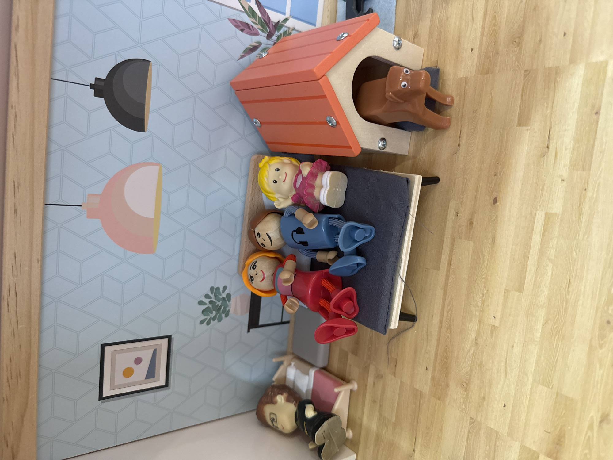 A miniature dollhouse scene is depicted, featuring a light blue wall with a geometric pattern and two hanging lamps, one pink and one black. On the wall, there is a framed picture with abstract shapes. In the foreground, a doll is lying in a small bed. To the right of the bed, three dolls are seated on a dark gray couch. The doll on the left is wearing a red outfit, the middle doll is in a blue outfit with headphones, and the doll on the right is wearing a pink dress. Next to the couch is a small, orange doghouse with a brown dog figure partially inside. The floor is a light brown, wood-grain texture. There are no visible elements that place this scene in Burgas, Bulgaria.