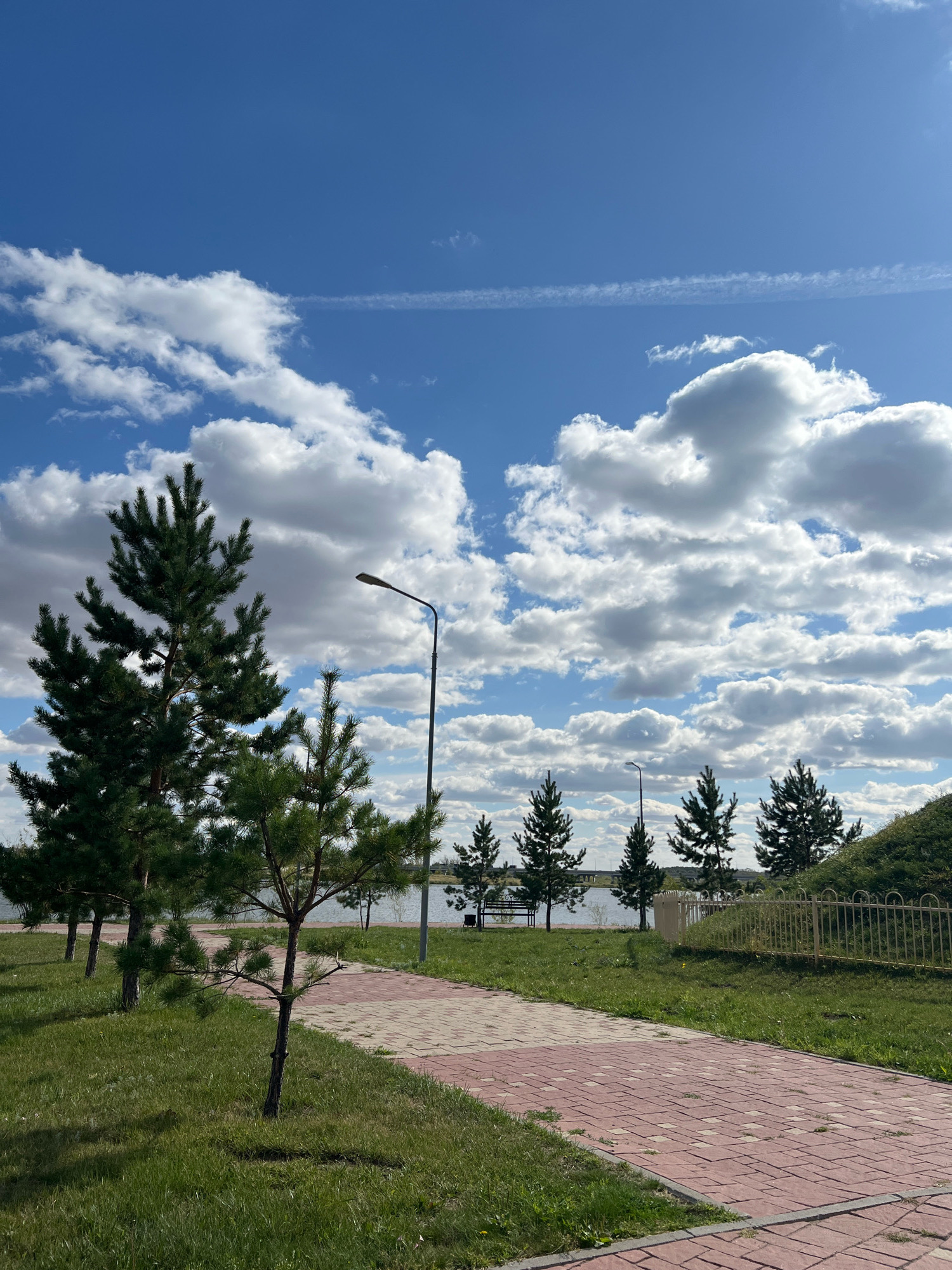 The image shows a paved walkway made of red and beige bricks, lined with green grass. Several pine trees of varying heights are planted along the walkway. A tall lamppost stands near the walkway. In the background, there is a body of water, likely a lake or river, with a bridge visible in the distance. The sky is blue with fluffy white clouds and a contrail from an aircraft. A metal fence runs along the right side of the image, separating the walkway from a grassy embankment. The scene depicts an outdoor recreational area in the city of Kostanay, Kazakhstan, possibly a park or embankment along the Tobol River.
