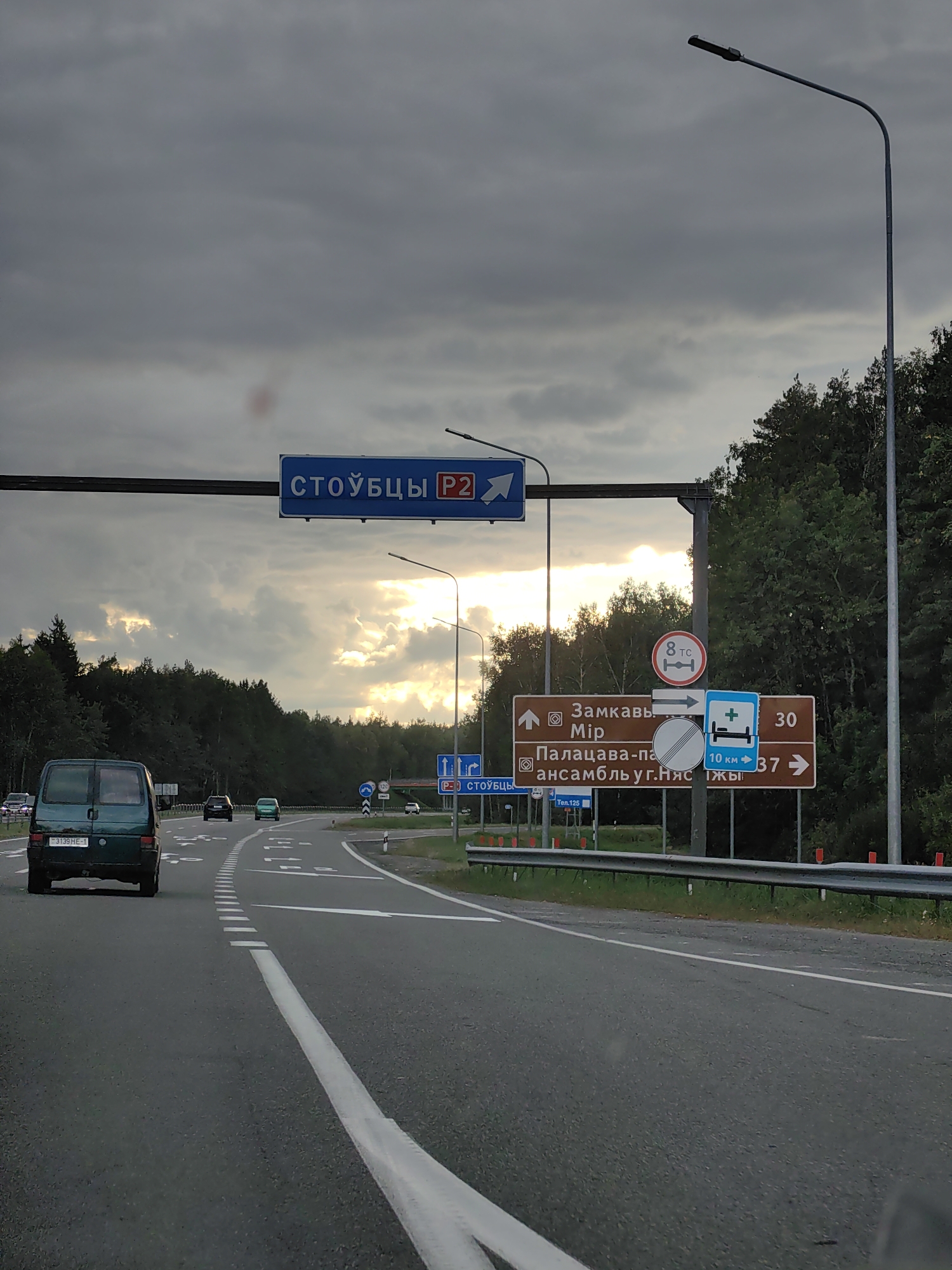 This image captures a view from a vehicle driving on a paved road in the vicinity of Заямнаўскі сельскі Савет, Belarus. The sky is overcast with dramatic clouds, and the sun is breaking through in the distance, casting a warm glow. The road is marked with white dashed lines, indicating multiple lanes. Several vehicles are visible on the road, including a dark green van in the foreground to the left, a black car behind it, and a green car further ahead. A collection of road signs are prominently displayed on the right side of the road. One sign indicates "СТОЙБЦЫ P2," a blue directional sign for a destination named Stoubtsy. Another set of brown signs directs to "Замкавы Мір" (Castle Mir) and "Палацава-паркавы ансамбль у г.Нясвіж" (Palace and Park Ensemble in Nesvizh), with arrows indicating directions and distances. There's also a blue sign with a medical cross and a bed symbol, suggesting a hospital or medical facility, with the text "10 км → 37 →". Additional smaller signs with numbers and symbols are also present. The road is bordered by dense green trees on both sides, with a metal guardrail running along the right side. The overall scene depicts a typical highway environment in a rural or semi-rural Belarusian setting.