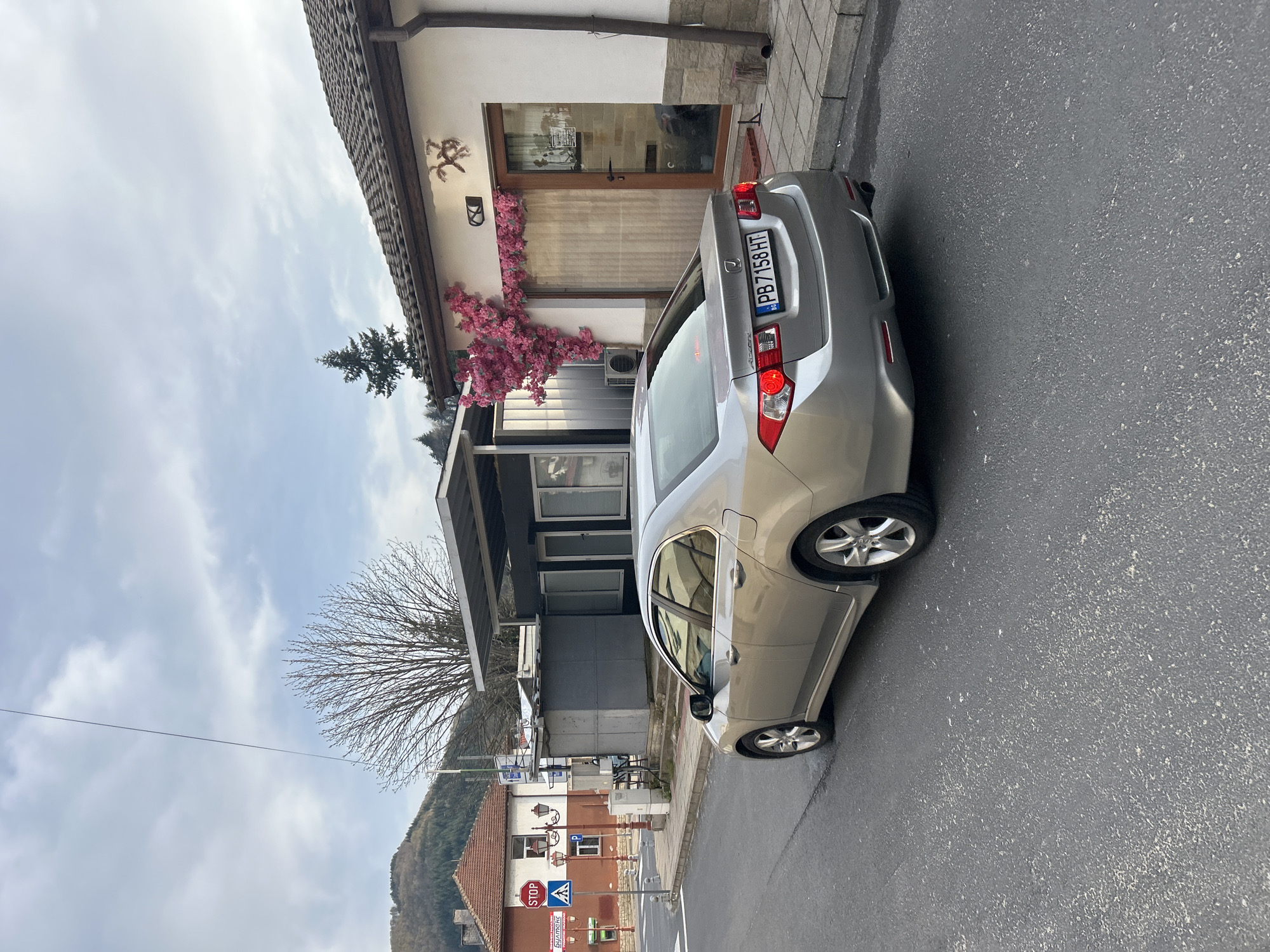 The image shows a silver Honda Accord parked on the side of a street in Zlatograd, Bulgaria. The car's license plate reads "PB 7158 HT." The vehicle is situated in front of a building adorned with pink flowers. A stop sign, crosswalk sign, and building are visible in the background, indicating an urban intersection. The sky is overcast.