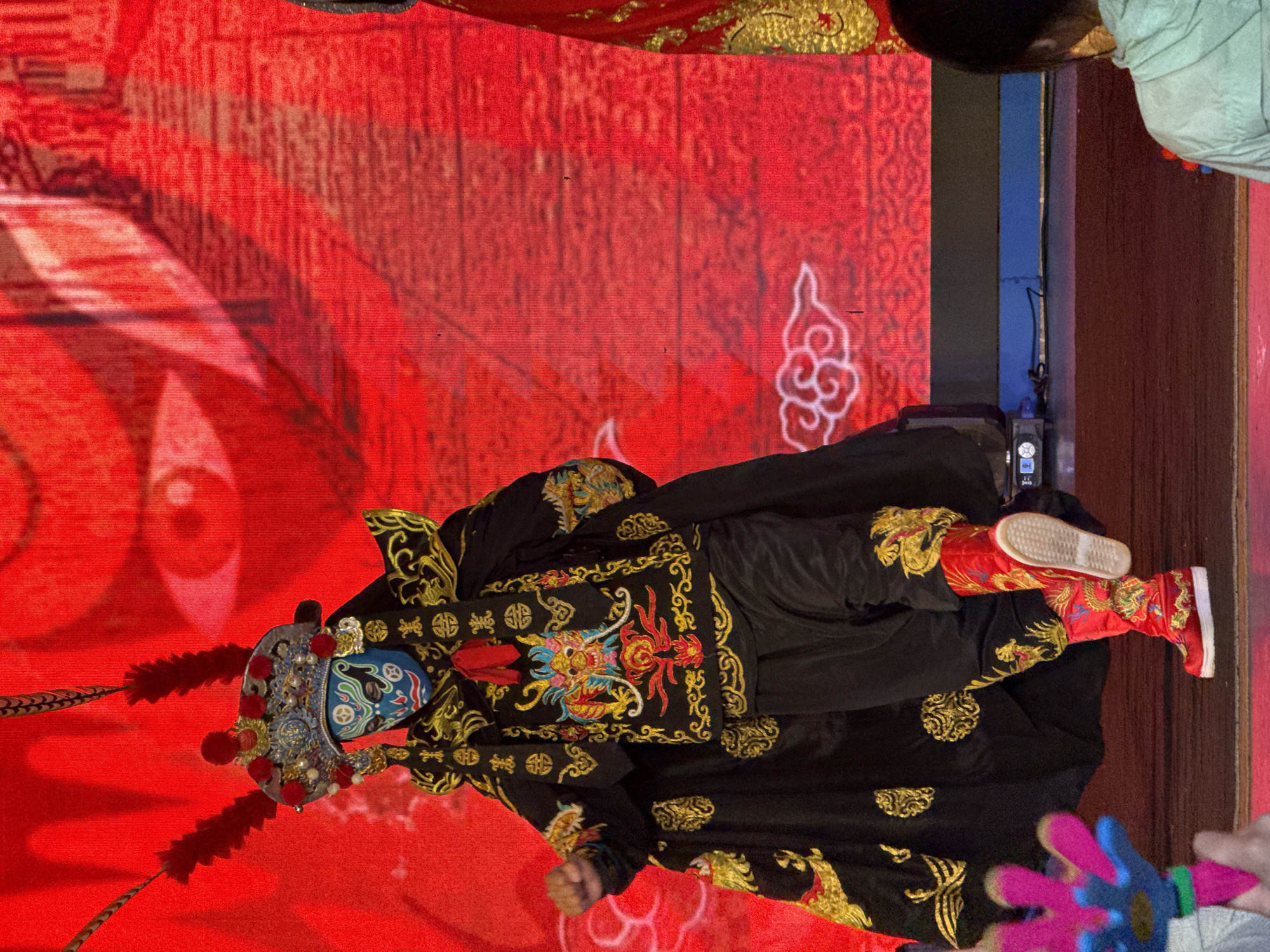 The image shows a performer in a Chinese opera costume, likely photographed in a theatrical or cultural setting. The performer wears a complex headdress with feathers and a painted facial mask in blue, red, and white tones. The costume is predominantly black with ornate gold embroidery, featuring dragon motifs and other decorative patterns. They also wear red boots with gold embroidery.
The backdrop is a vibrant red, with a large eye or face design on one side and cloud-like patterns. The image's orientation suggests it was either taken vertically or has been rotated.
Given the details and the request, it's reasonable to infer that the image was likely taken in Yuzhong District, China, specifically at a cultural performance or event showcasing traditional Chinese opera.