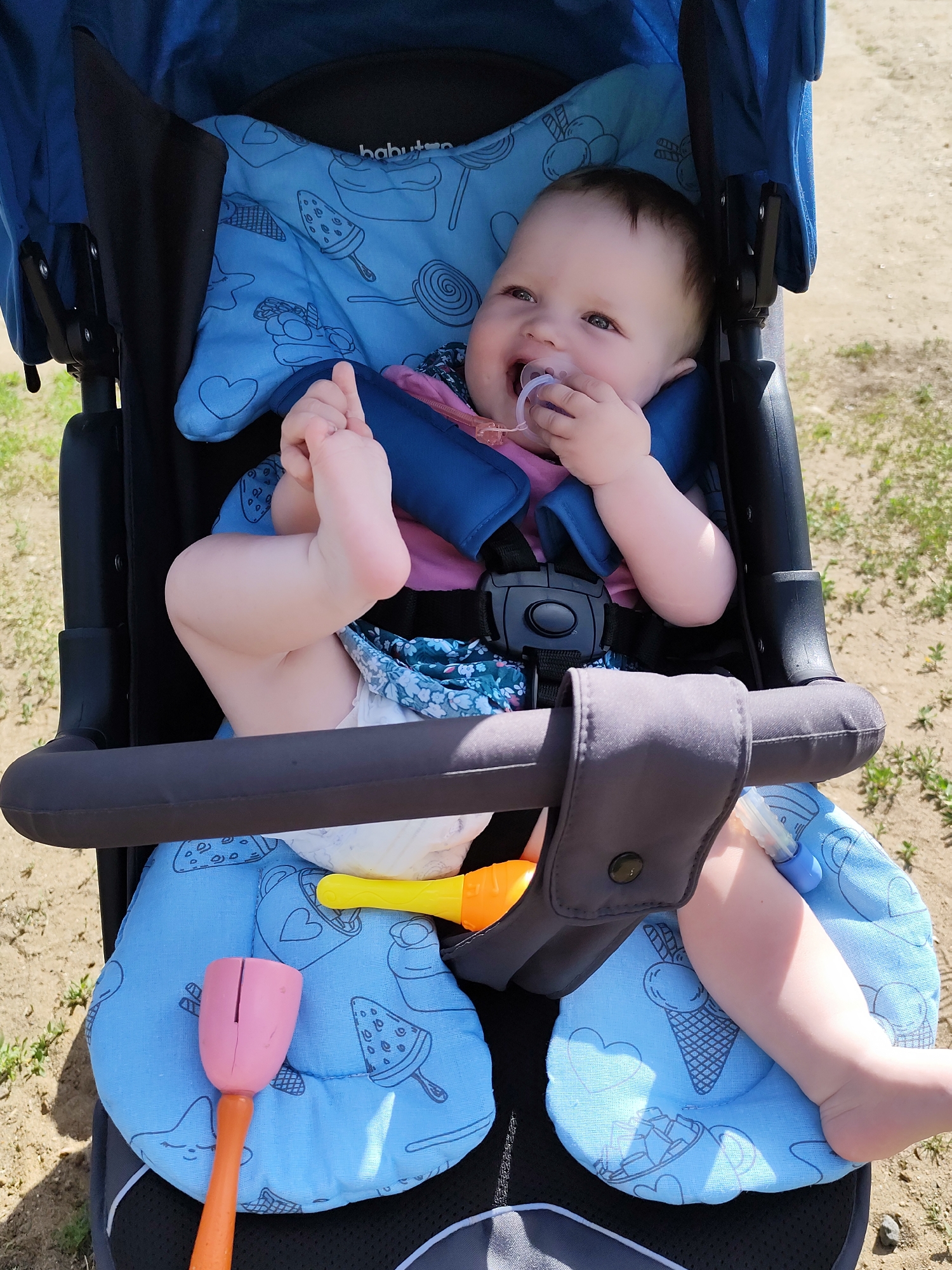 A baby is sitting in a blue stroller. The baby is wearing a diaper and a blue floral dress with a pink shirt. The baby has a pacifier in their mouth and is holding their foot with their hand. There are two toys in the stroller. The stroller is on a dirt path with grass growing on either side. The location is possibly in the city of Фоки, Russia.