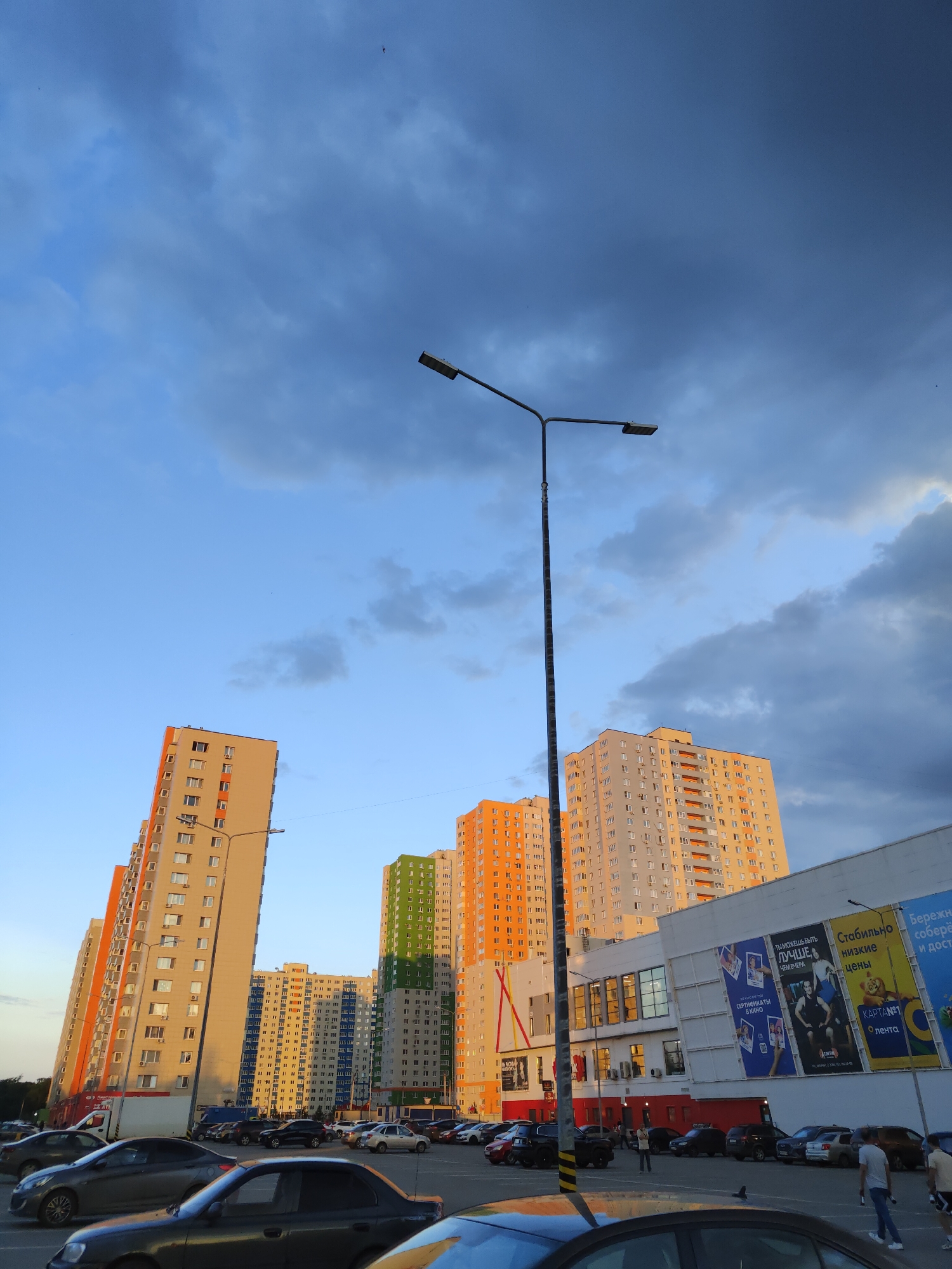 The image depicts a wide shot of a street scene in Ufa, Russia. In the background, several modern, high-rise apartment buildings with brightly colored facades (orange, green, and beige) dominate the skyline. Some of these buildings are partially illuminated by the setting sun. In the mid-ground, a large, white commercial building with signage is visible, featuring advertisements with text in Russian. A tall street lamp stands prominently in the foreground, casting a shadow. The foreground and mid-ground are filled with numerous parked cars of various makes and models, suggesting a parking lot or a street with ample parking. A few individuals are visible on foot in the parking area, some walking and one person appears to be holding a microphone or a similar object. The sky above is a mix of blue with scattered, dark clouds. The overall scene suggests an urban environment during the late afternoon or early evening.