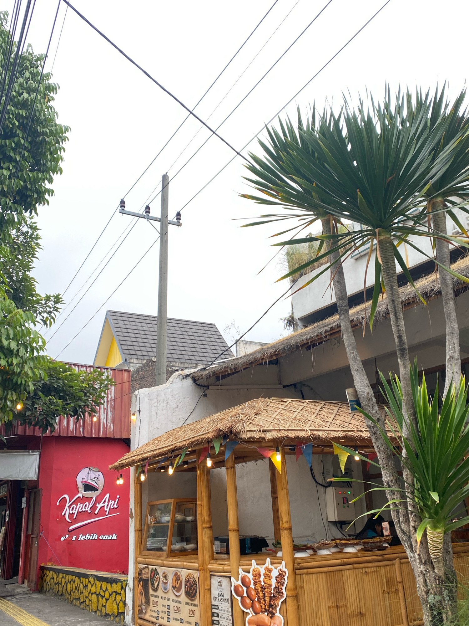 An exterior, eye-level, slightly low-angle medium shot captures a street scene in an outdoor setting. The image is dominated by a bright red building with white lettering that reads "Kapal Api" and "e's lebih enak" in a stylized font, with a silhouette of a ship above the text. To the right of the red building, a small, open-air food stall with a thatched roof and bamboo construction is visible. The stall displays food items and menus, with a few decorative flags strung across its top. Palm trees and other lush green foliage are present in the foreground and to the left, adding a tropical feel. Overhead, utility poles and electrical wires crisscross the sky, which is overcast and appears to be a bright white. The ground in front of the red building is a patterned sidewalk with yellow and white accents. The overall scene suggests a street-side vendor or cafe in a warm climate.