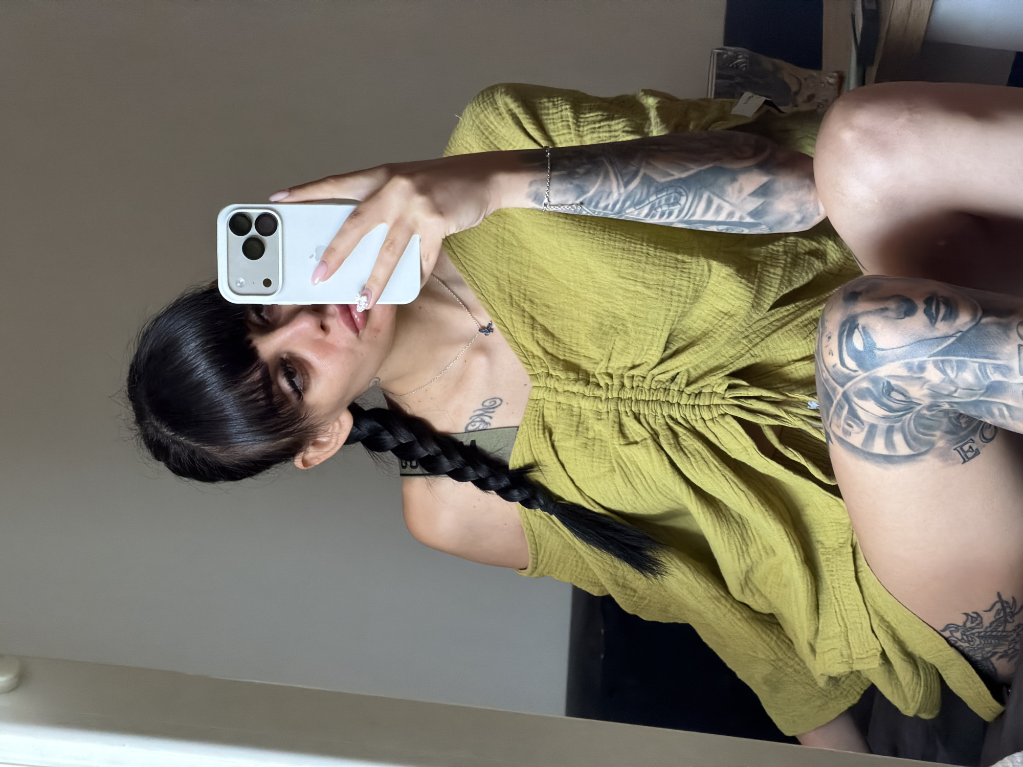 A young woman with dark hair in a braid and bangs is taking a mirror selfie. She is wearing a green off-the-shoulder top and has extensive tattoos on her arms and thighs. She is holding a white smartphone in her right hand. The background appears to be an indoor setting with neutral-colored walls. There is no indication of the city of Plovdiv, Bulgaria, in the image.