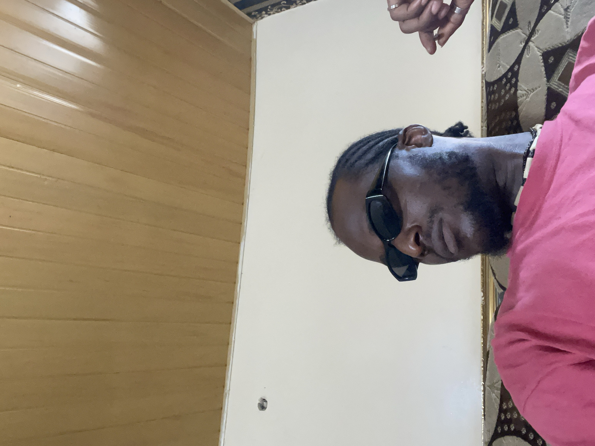An adult male, with dark skin, braided hair, a short beard, and mustache, is depicted from the chest up, seated indoors. He wears rectangular black sunglasses, a solid pink t-shirt, and a black and white beaded necklace. Multiple rings adorn his right hand, which is slightly raised with fingers curved. He is looking downwards and to his left. The background features a light brown, vertically grooved ceiling with visible light reflections. Beneath the ceiling is an off-white wall, presenting a dark circular indentation on the upper left side. In the upper right corner, a section of dark, patterned wallpaper is visible. The immediate foreground shows the intricate, dark-patterned upholstery of a sofa or cushion, topped with a golden trim.