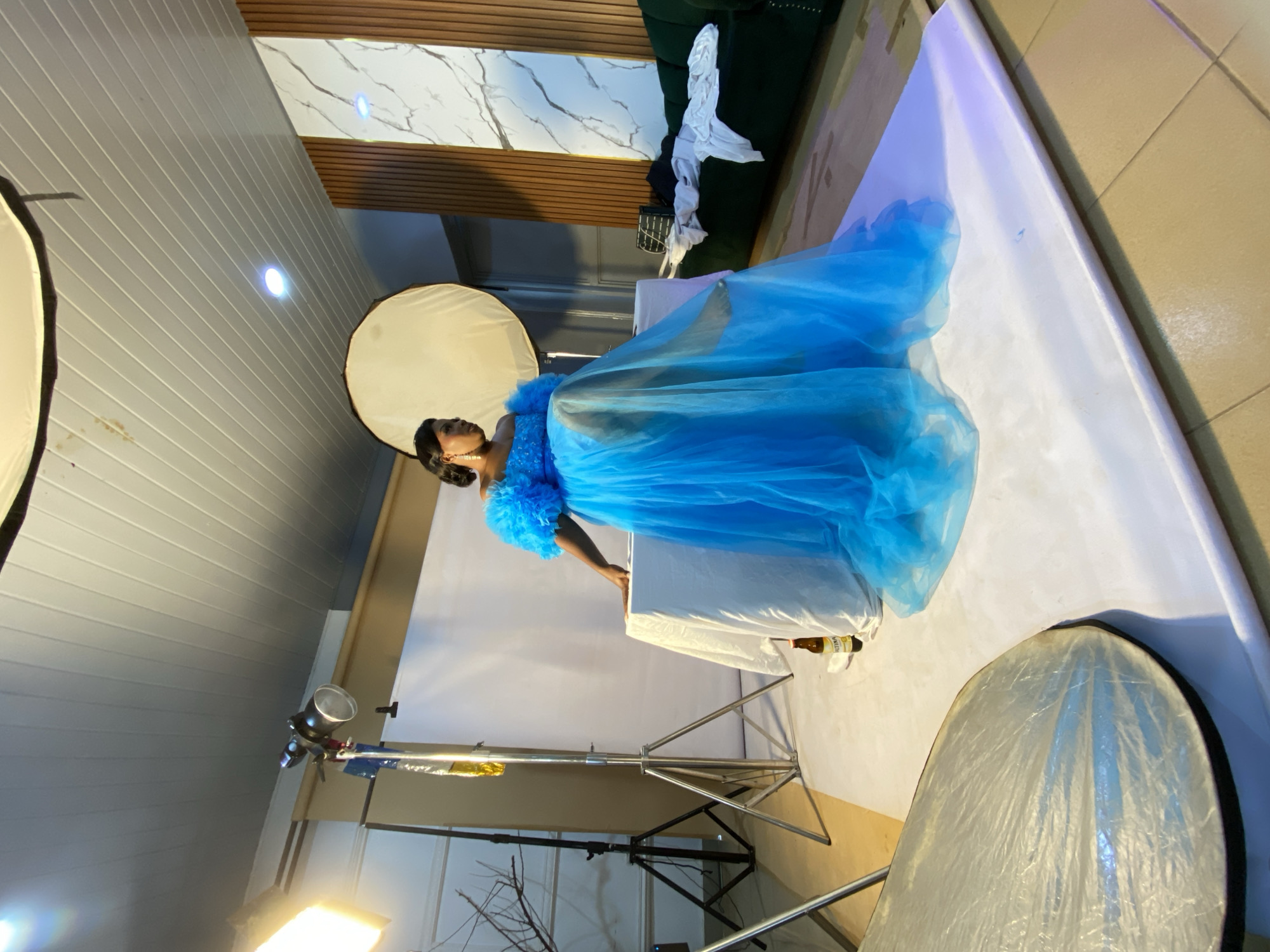 A woman in a flowing blue gown poses in a studio setting. The gown is voluminous and made of a sheer fabric, with ruffled detailing on the shoulders. The woman is facing to her left, with her head turned slightly towards the camera. She has dark hair pulled back and appears to be wearing earrings. She is standing behind a white surface, which is part of the studio setup.
The studio is equipped with professional lighting equipment, including softboxes and stands, indicating a photoshoot is in progress. In the background, there is a dark green sofa with some white fabric draped over it, and a wall with a marble-like pattern and wooden slats. The floor appears to be tiled. A bottle with a label is visible on the white surface near the woman's gown. The overall impression is that of a formal portrait session. The lighting suggests it is indoors, likely during the day, but the specific time of day cannot be determined.