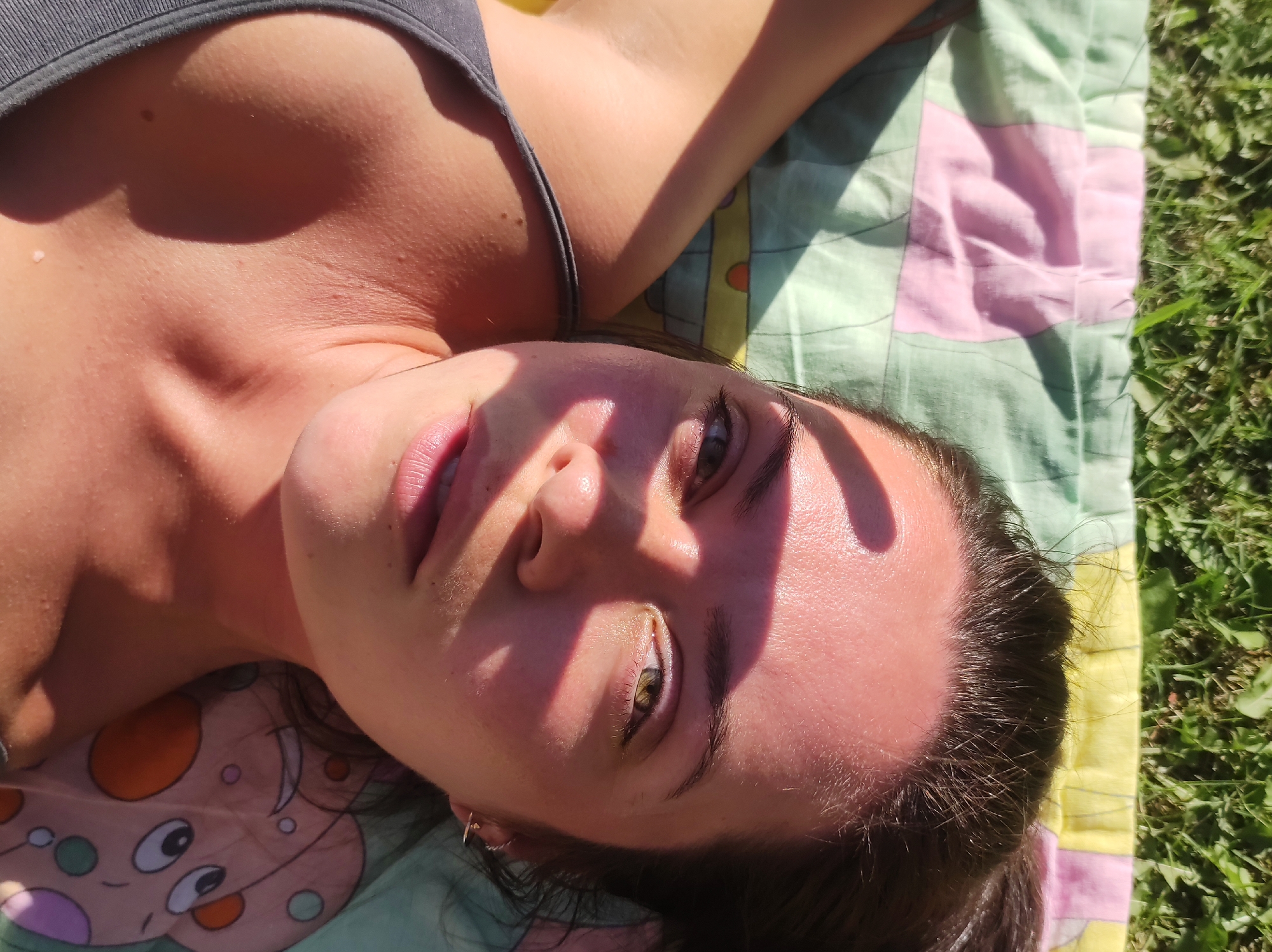 The image shows a close-up, overhead view of a young person lying on a blanket outdoors. The person is facing upwards, with their head tilted. Sunlight casts shadows across their face. They are wearing a gray tank top. The blanket is patterned with squares of pink and green, and there are some cartoonish figures printed on it. The blanket is laid out on green grass. The scene appears to be in a natural setting, possibly a park or backyard. There is no visible indication of the location being in the city of Фоки, Russia.