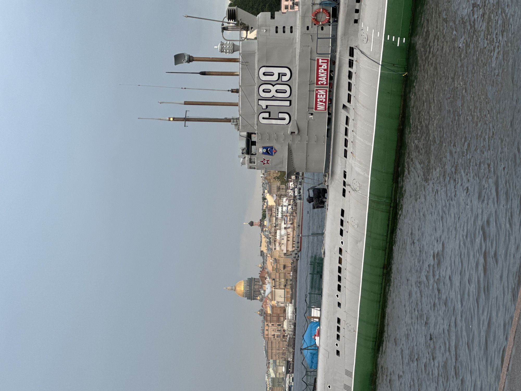 The image shows a grey submarine with a green waterline, identified as "C189" and bearing the Russian text "МУЗЕЙ ЗАКРЫТ" (Museum Closed). The submarine is moored in the Neva River in Saint Petersburg, Russia, with a cityscape of historical buildings in the background. Most prominent among the buildings is the golden-domed Saint Isaac's Cathedral. The sky is overcast, and the water appears dark and choppy. The scene suggests a somber or quiet day, fitting with the "Museum Closed" sign on the submarine. There are no people visible in the image.