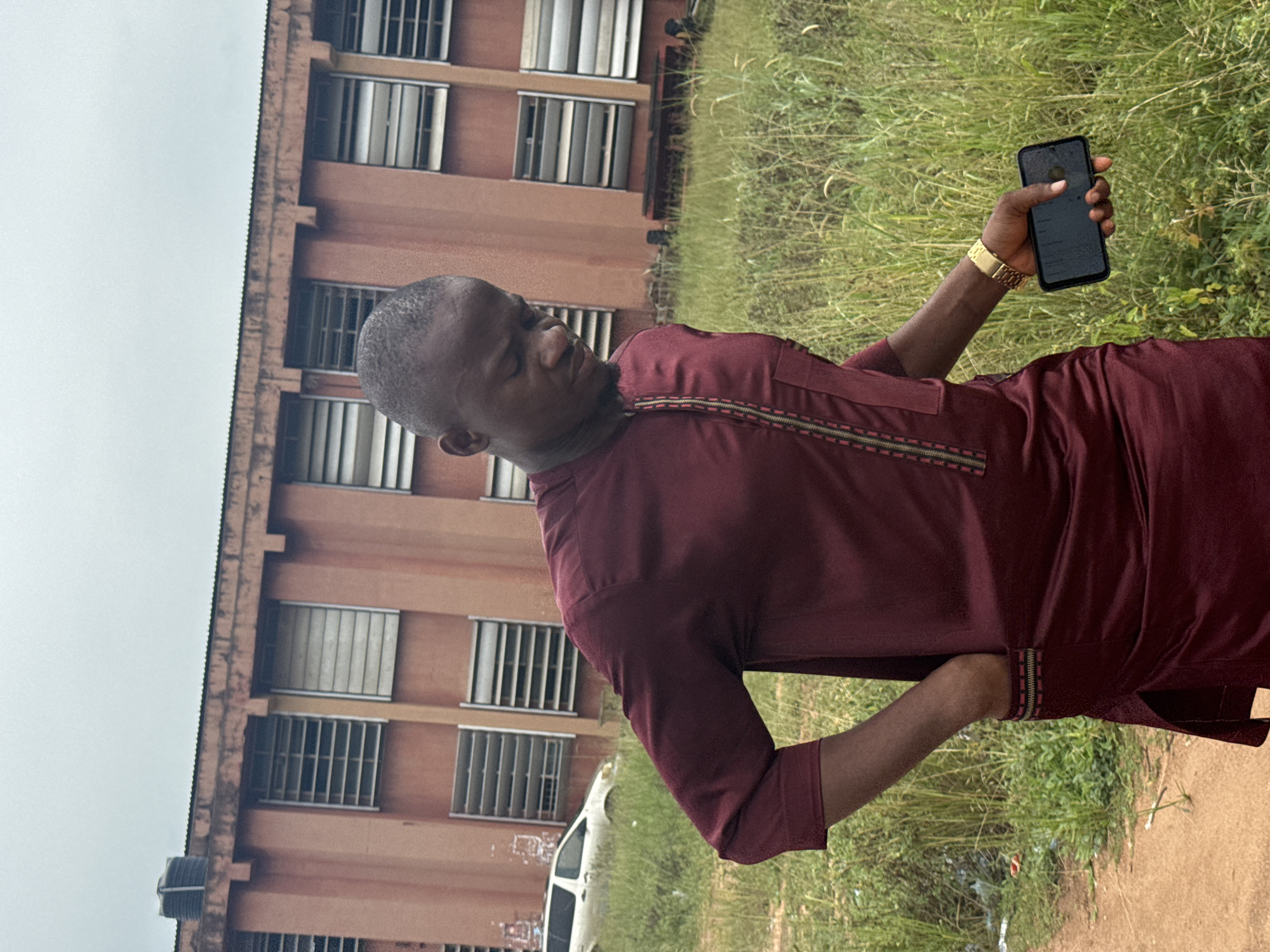An outdoor, vertically oriented image, originally rotated 90 degrees counter-clockwise, depicts a man standing in front of a building with a grassy area. The man, who is Black/African with a closely shaved head and light facial hair, is wearing a dark red/maroon traditional-style tunic featuring a zippered neckline and patterned trim. He stands with his left hand on his hip and his right arm extended, holding a black smartphone. A gold-colored wristwatch is visible on his right wrist. He looks downwards with a neutral expression.
The setting includes a multi-story building, painted in an orange-brown/ochre color, with numerous windows fitted with light-colored vertical blinds or louvers. The building appears somewhat weathered. A black cylindrical object, possibly a water tank, is visible on its roofline. A white vehicle, potentially a van or SUV, is partially seen on the grassy ground near the building. To the man's right and in the background is an expanse of green grass mixed with taller, wilder vegetation. The sky is overcast and uniformly light grey, suggesting a cloudy daytime. The scene evokes a casual moment in Ihiagwa, Nigeria. There is no discernible text visible in the image.