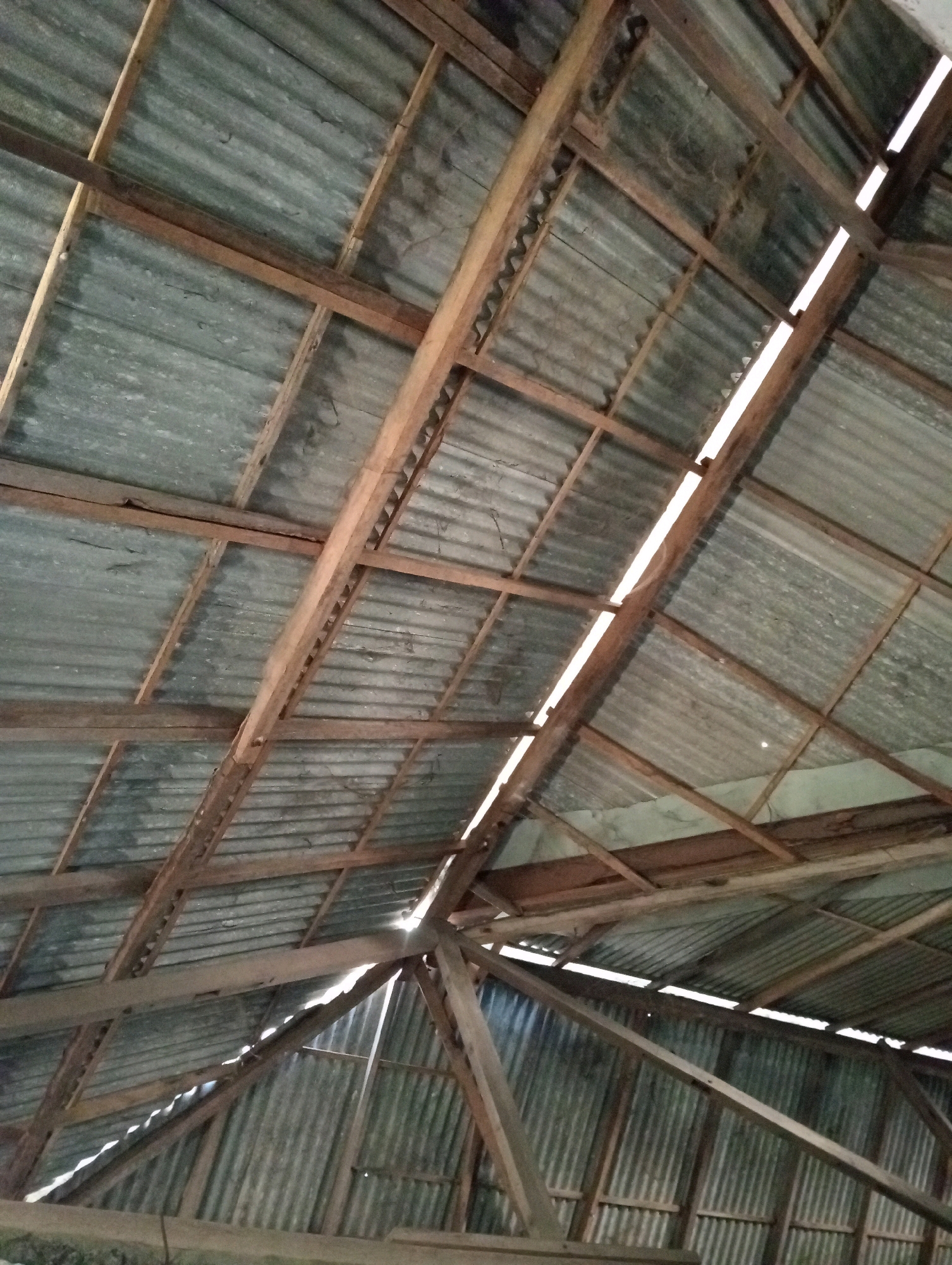 An interior view of a corrugated metal roof supported by a wooden beam structure is shown. The roof slopes upwards from bottom to top and right to left. Gaps in the roof allow light to enter, creating bright linear highlights. The wooden beams are weathered and show signs of construction. The location is identified as Pagerungan Kecil, Indonesia.